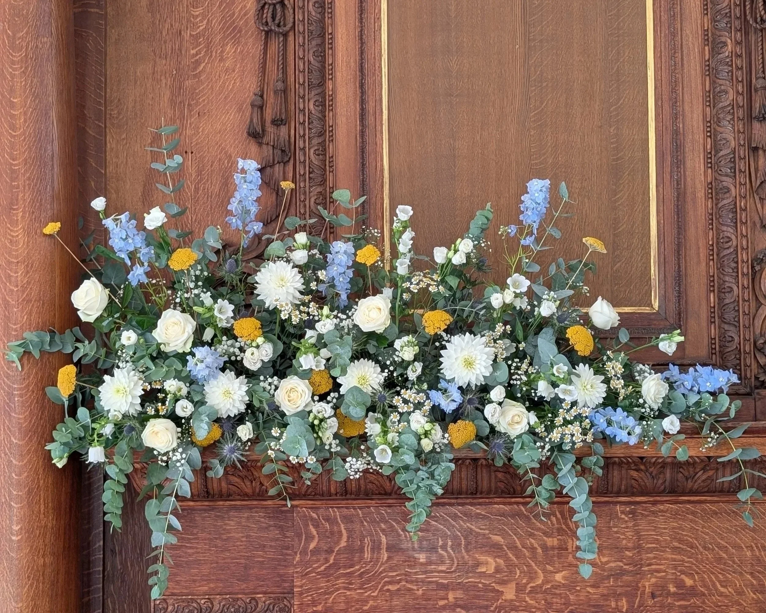 A floral arrangement with white roses, blue delphiniums, and yellow and white daisies in front of a carved wooden fireplace mantlepiece.