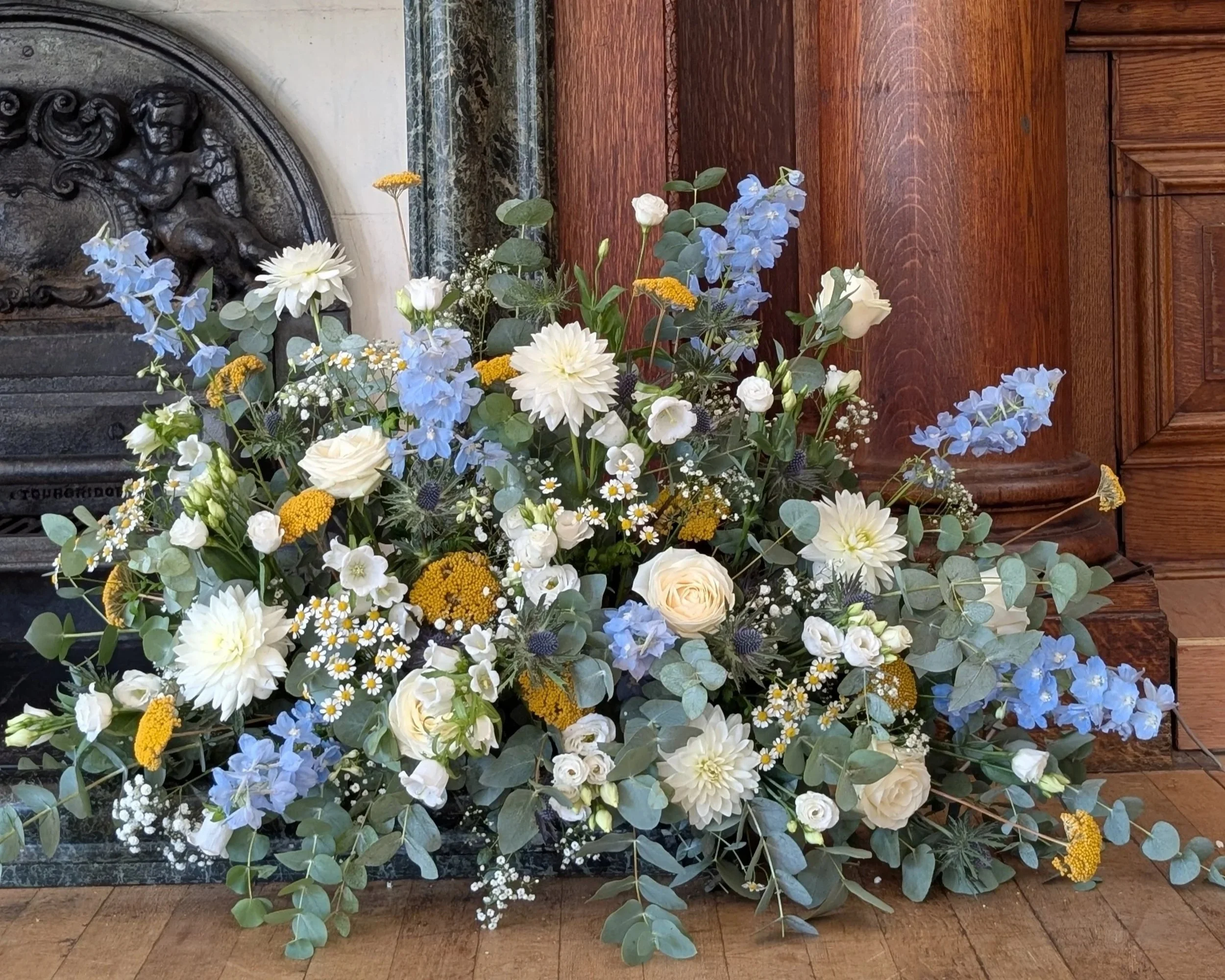 A flower arrangement with white dahlias, blue delphiniums, small white daisies, and greenery placed in front of a wooden panel and a decorative metal angel sculpture on the wall.