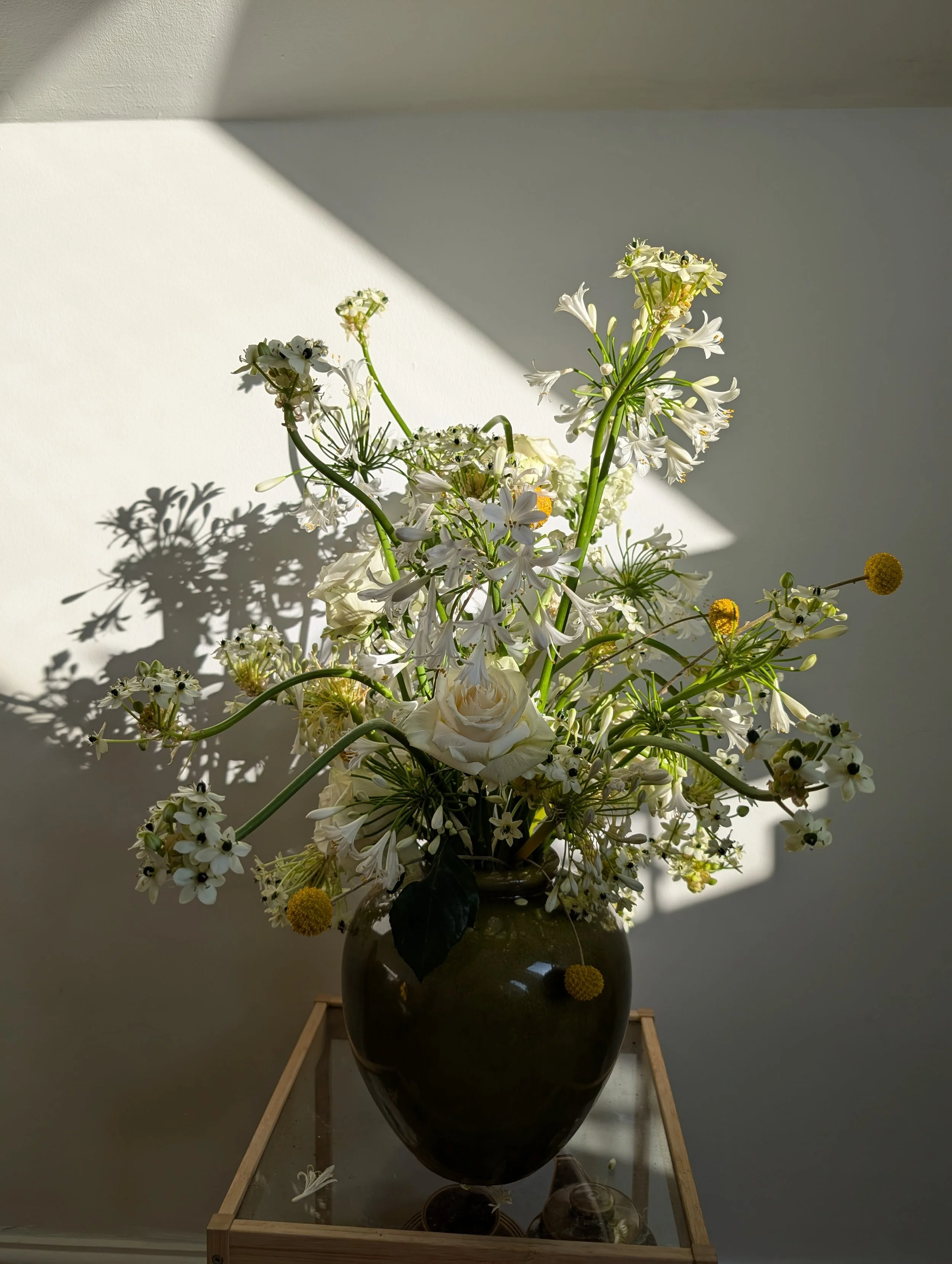 A dark brown ceramic vase filled with white and yellow flowers, casting shadows on a plain wall behind it, placed on a glass-topped wooden table.