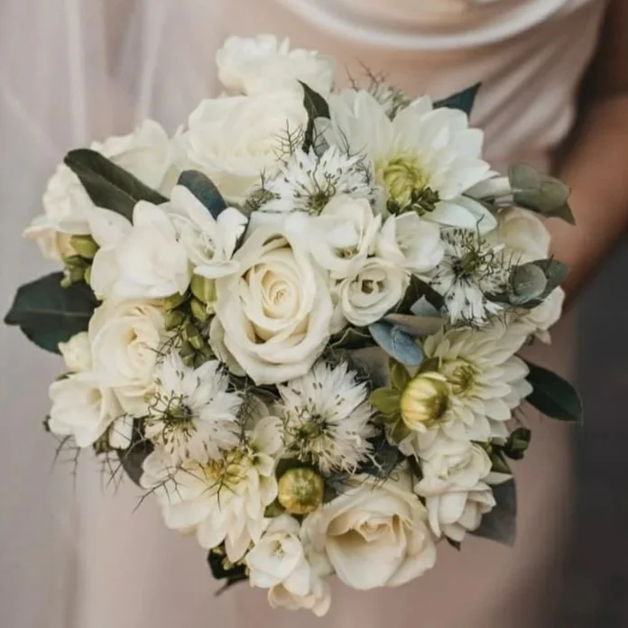 A bridal bouquet of white roses, dahlias, and other white flowers with green leaves and small decorative elements.