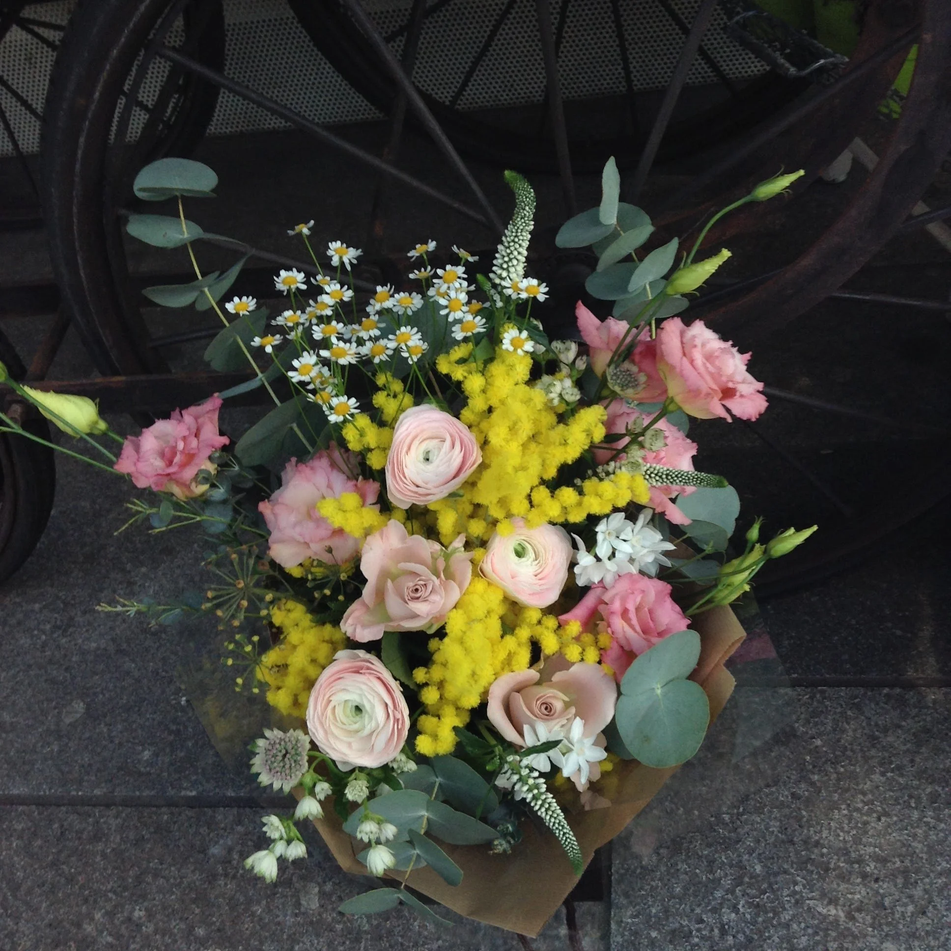 A bouquet of pink, white, and yellow flowers including daisies and lisianthus, with greenery such as eucalyptus, arranged on a brown paper wrap in front of a dark wicker chair.