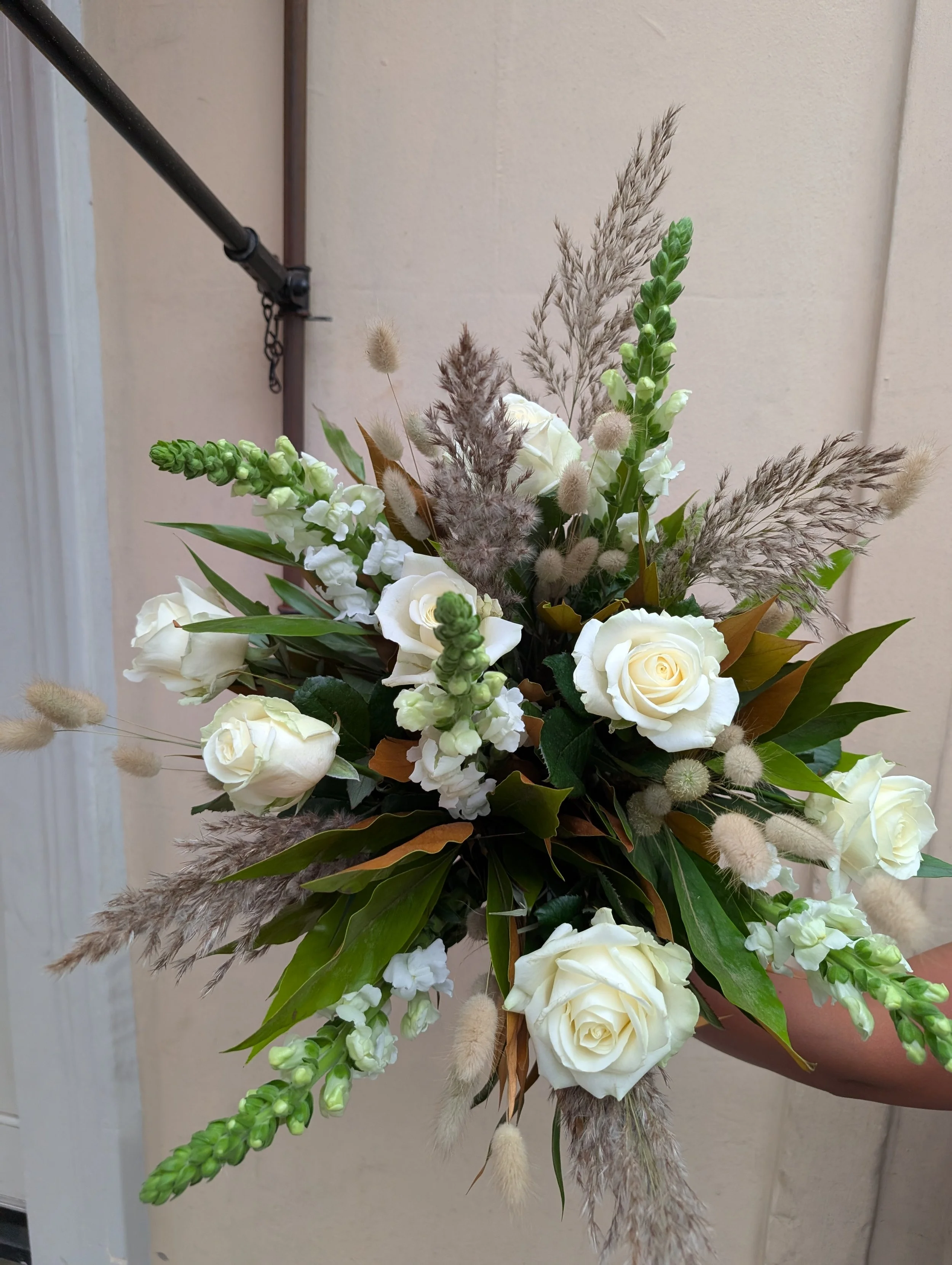 Arrangement of white roses, green buds, dried grasses, and foliage in a bouquet held by a person against a neutral wall background.