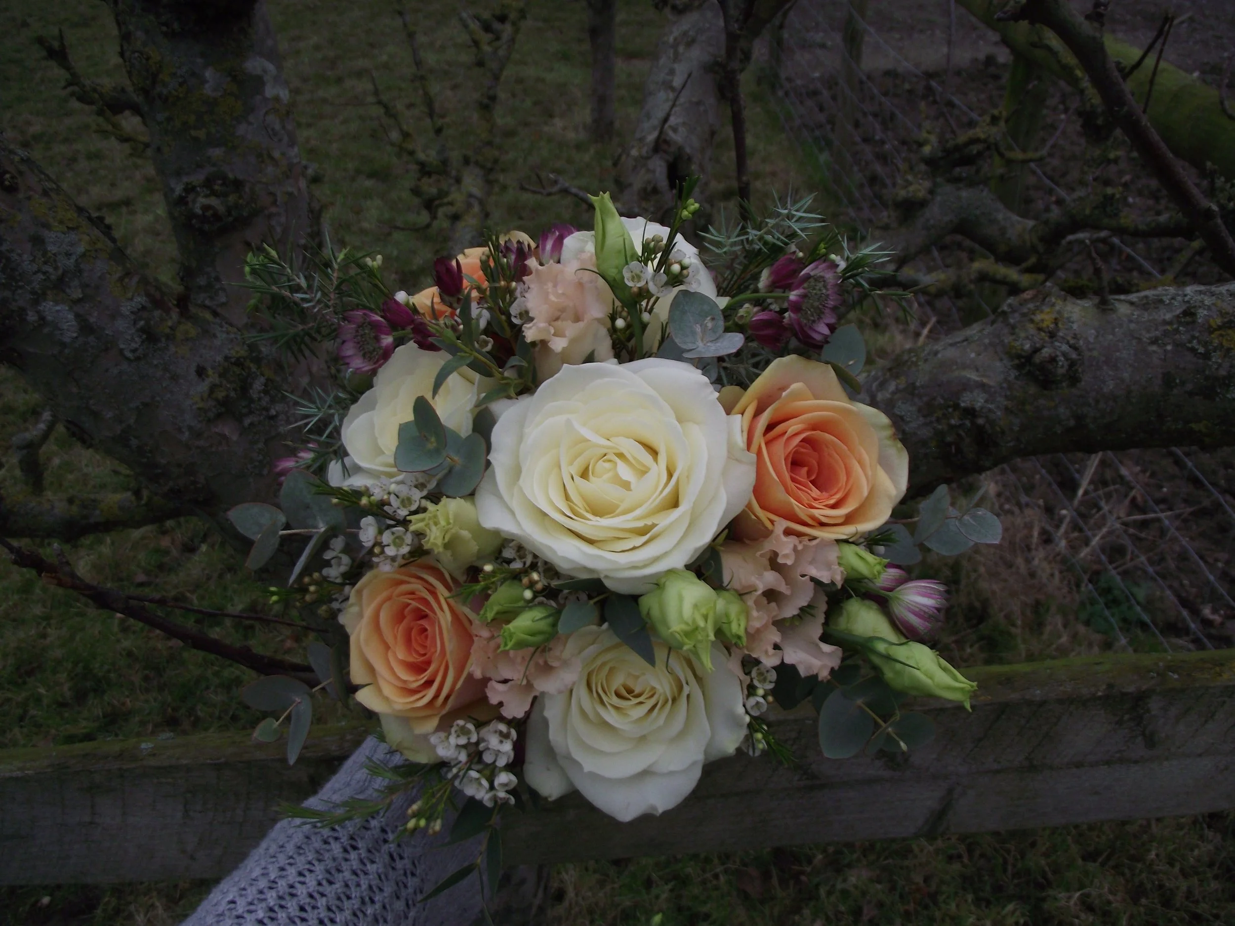 A bouquet of flowers with white, peach, and purple roses, green foliage, and small white flowers, held with a gray lace ribbon, against a dark outdoor background.