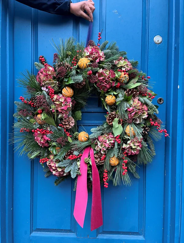 A colorful holiday wreath hanging on a blue door, decorated with pink hydrangeas, pinecones, tiny pumpkins, red berries, and a pink and red ribbon.