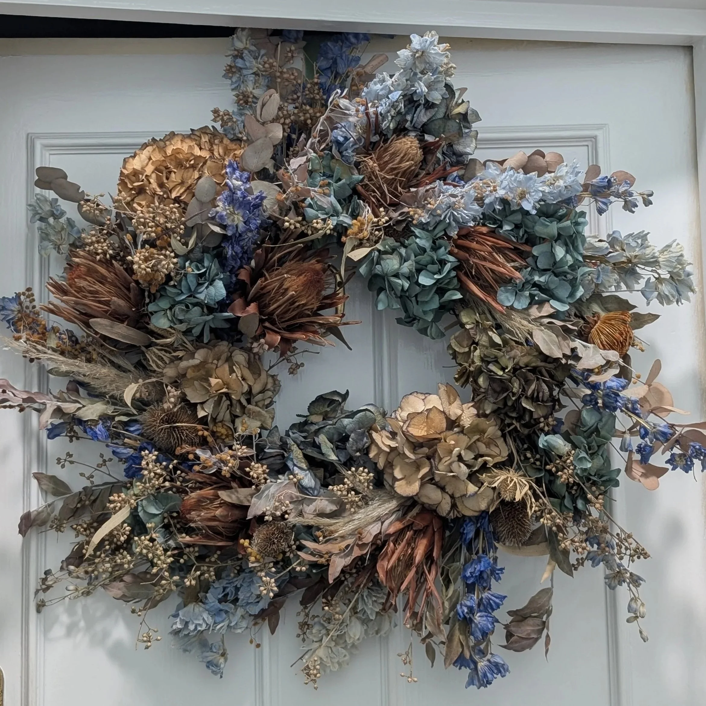Dried floral arrangement with blue, beige, and brown tones hung on a white wall.