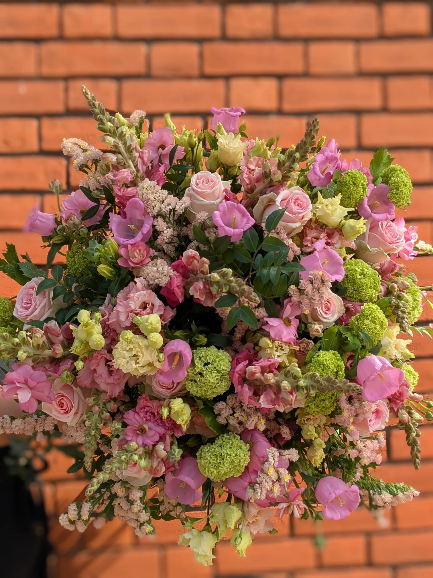 A large bouquet of mixed pink, white, and green flowers against a brick wall background.