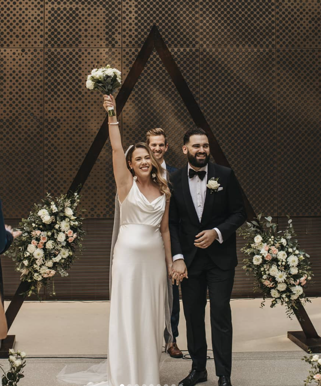 Bride and groom celebrating wedding, holding hands, with the bride raising a bouquet in the air, standing in front of a decorative geometric backdrop with floral arrangements.
