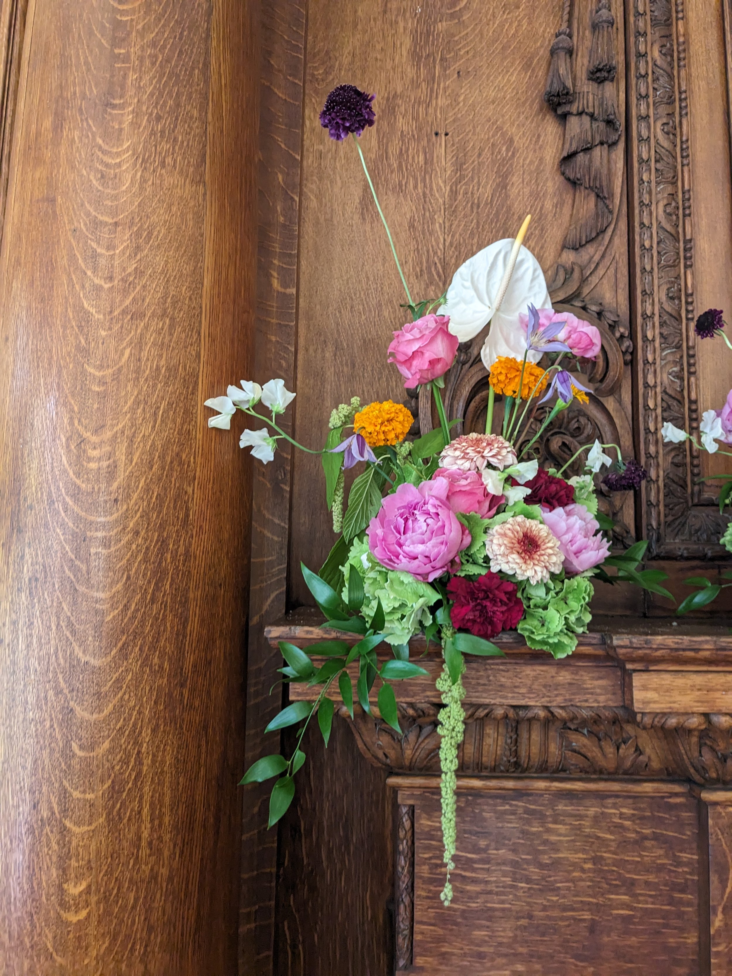 A floral arrangement on a wooden surface, featuring pink, purple, white, orange, and green flowers with green foliage.