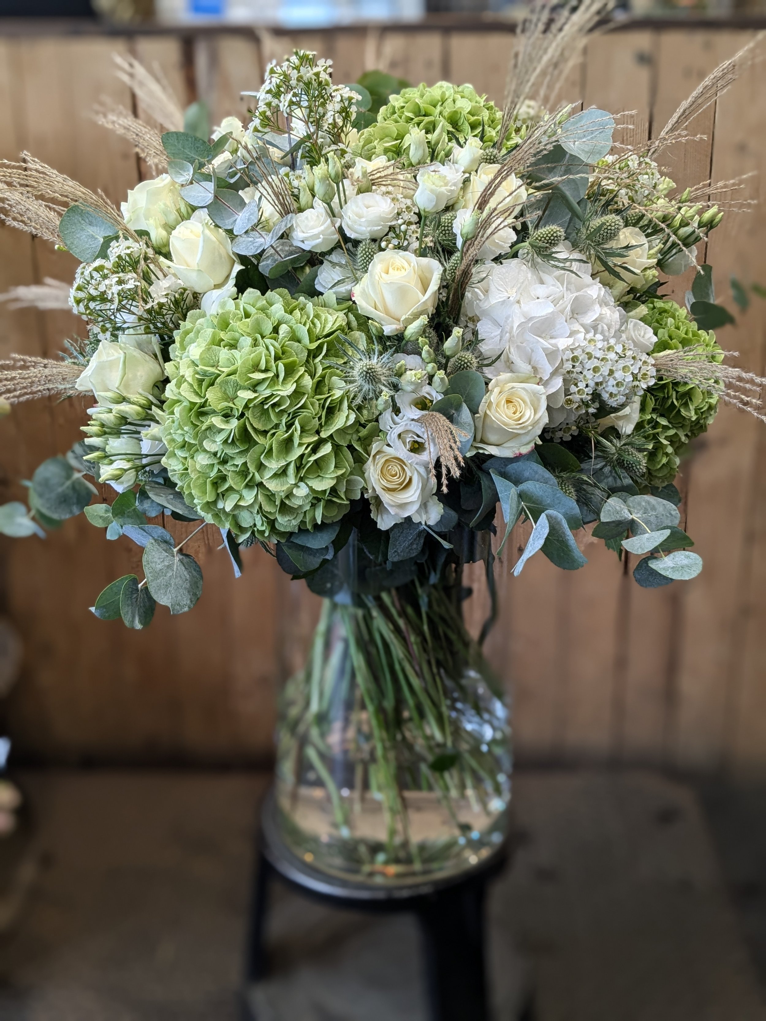 A bouquet of white roses, green hydrangeas, and assorted greenery in a clear glass vase on a black table.