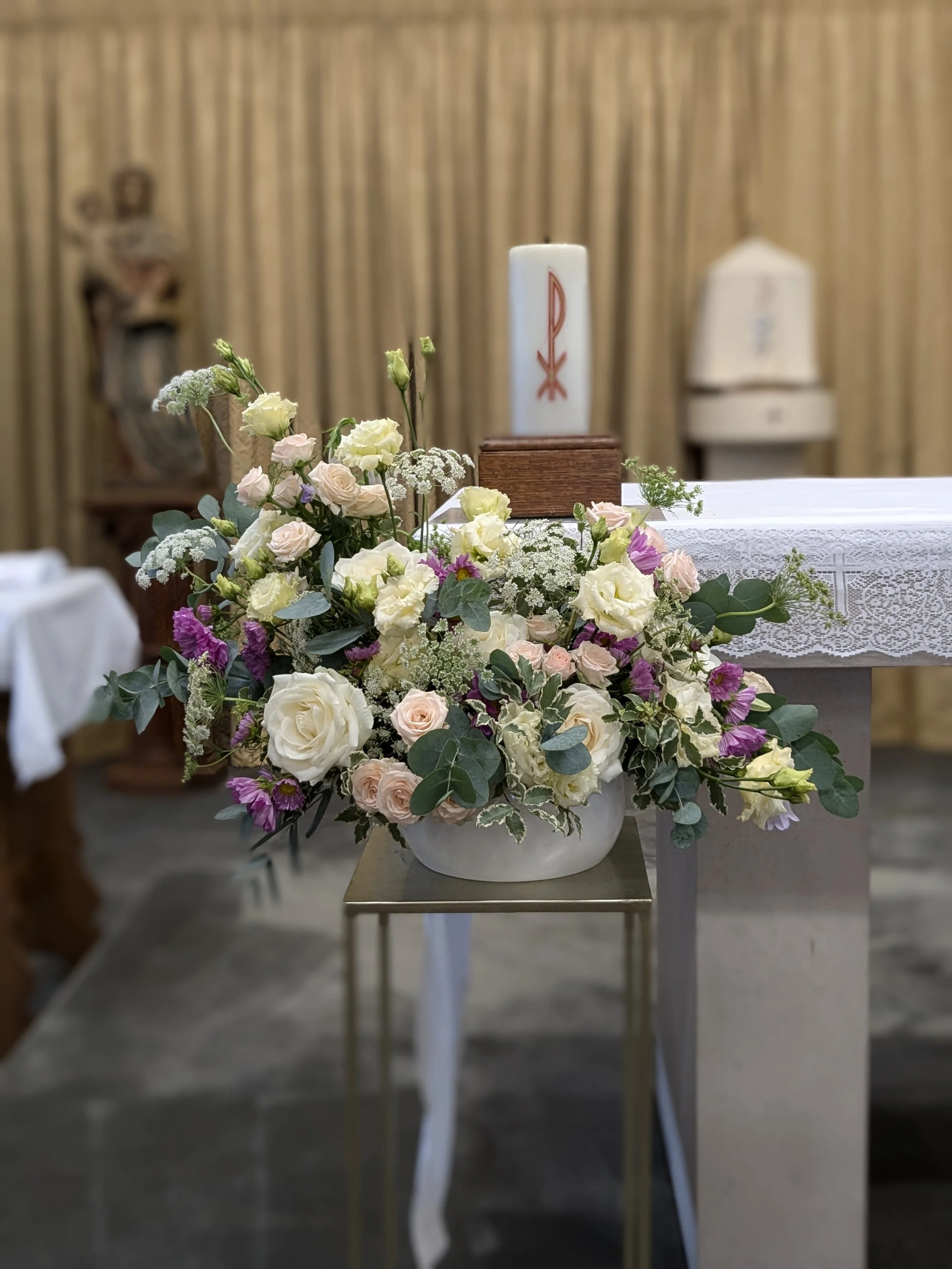 A floral arrangement with white, pink, and purple flowers, set on a table with a white lace cloth, in front of a religious altar with candles, in a church setting.