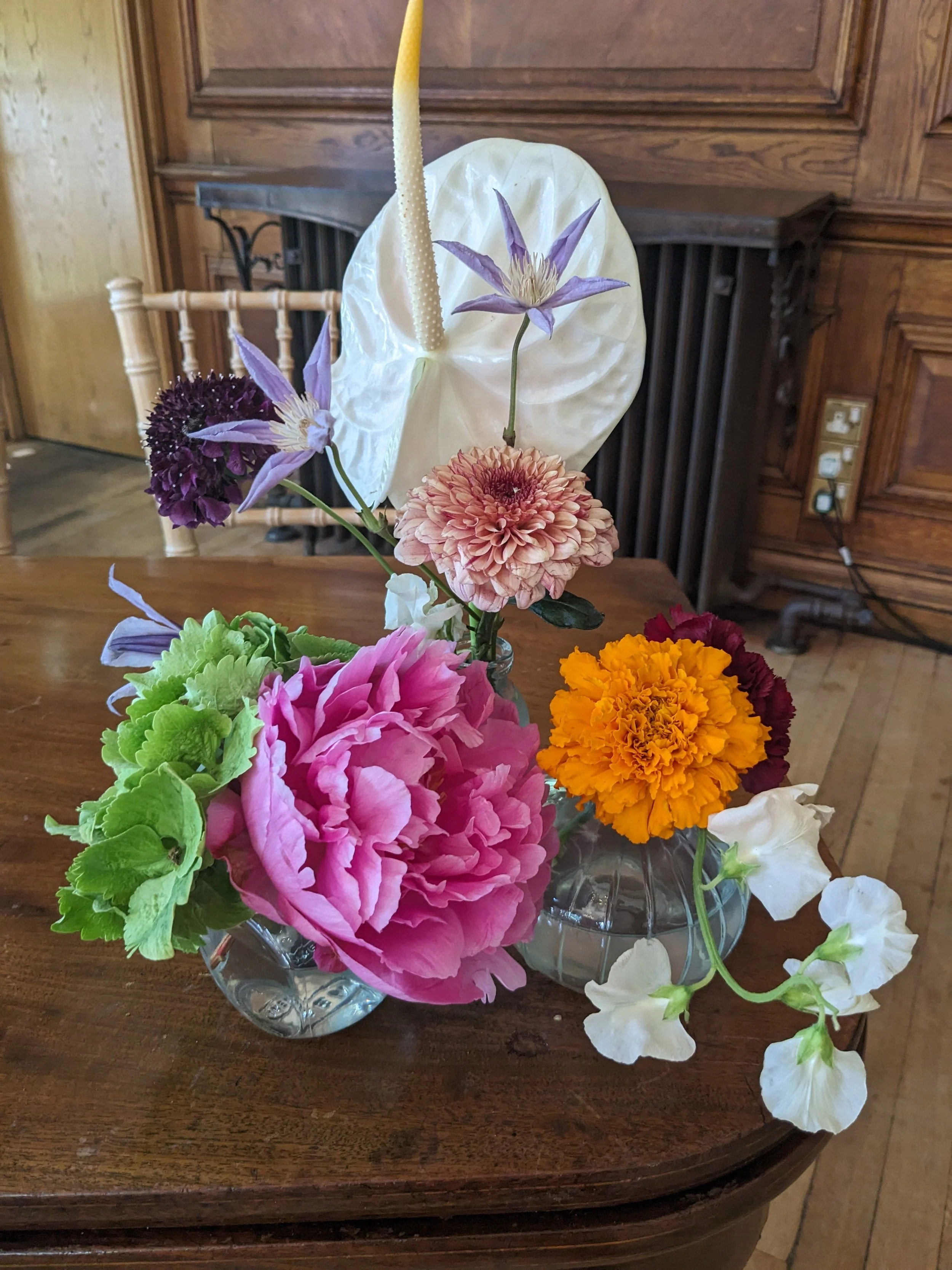 A glass bowl filled with colorful artificial flowers on a wooden table, with a wooden wall and electrical outlet in the background.