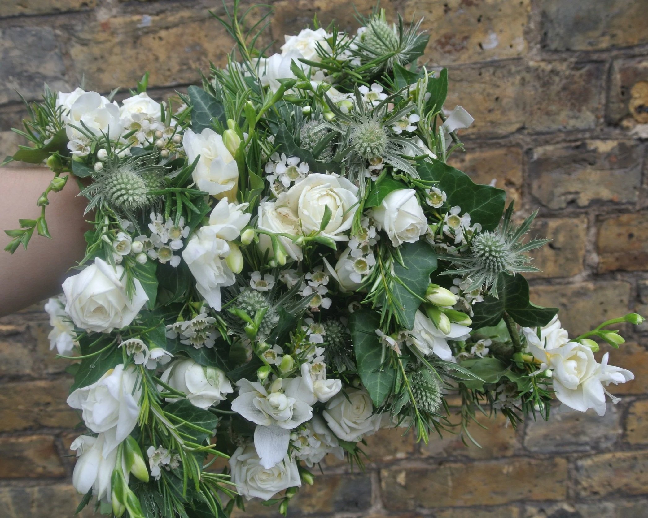 A bouquet of white flowers including roses, small white filler flowers, green leaves, and textured green spikes, set against a brick wall background.