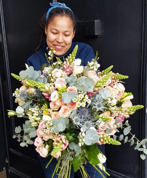 Black London Woman with a blue headband holding a large bouquet of pink, white, and green flowers, smiling in front of a black wall.