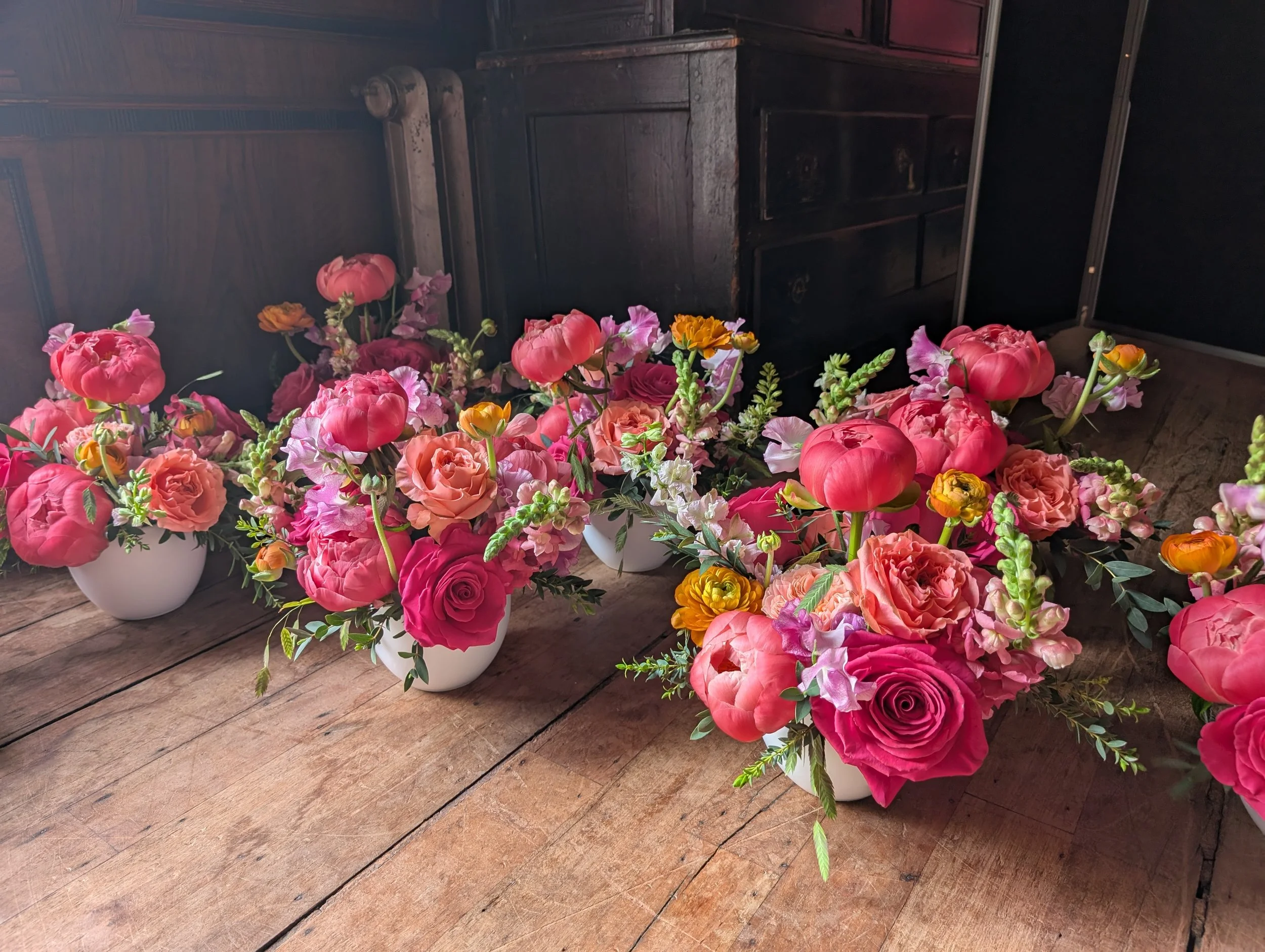 Multiple pink, red, orange, and purple flowers arranged in white bowls and placed on a wooden surface with a dark wooden cabinet or chest in the background.