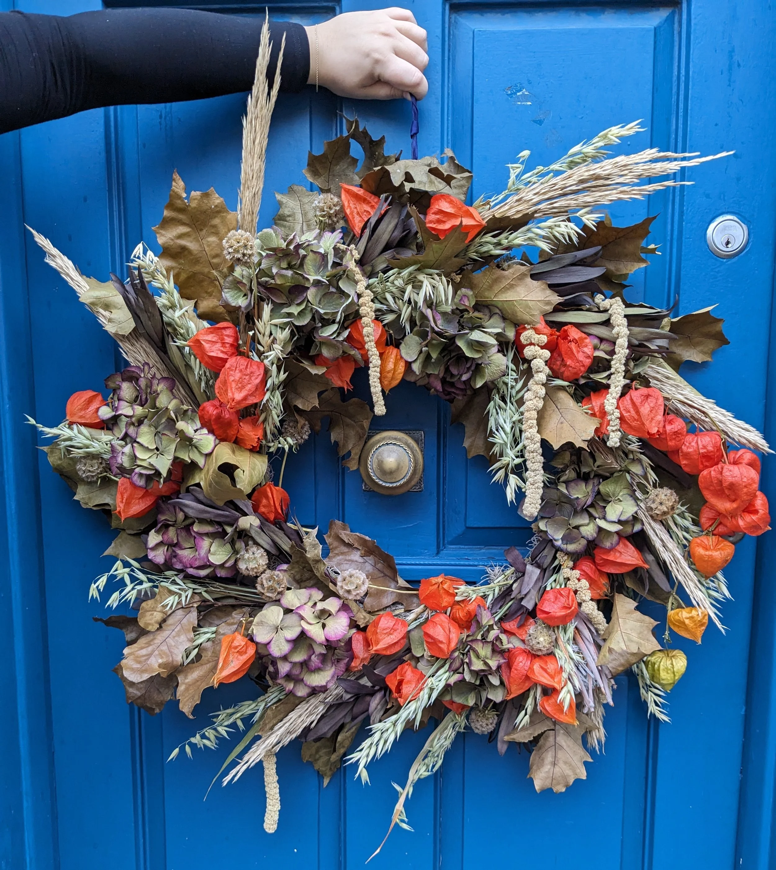 Close-up of a decorative autumn wreath hanging on a bright blue door. The wreath is made of various dried leaves, grasses, and orange paper flowers, with a small hand holding the purple string hanger at the top.