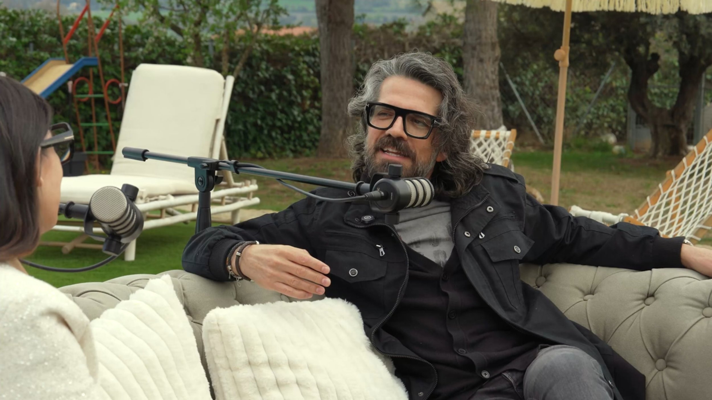 Hombre con barba y gafas negras hablando en grabadora mientras está sentado en un sillón en un jardín con juegos y árboles de fondo