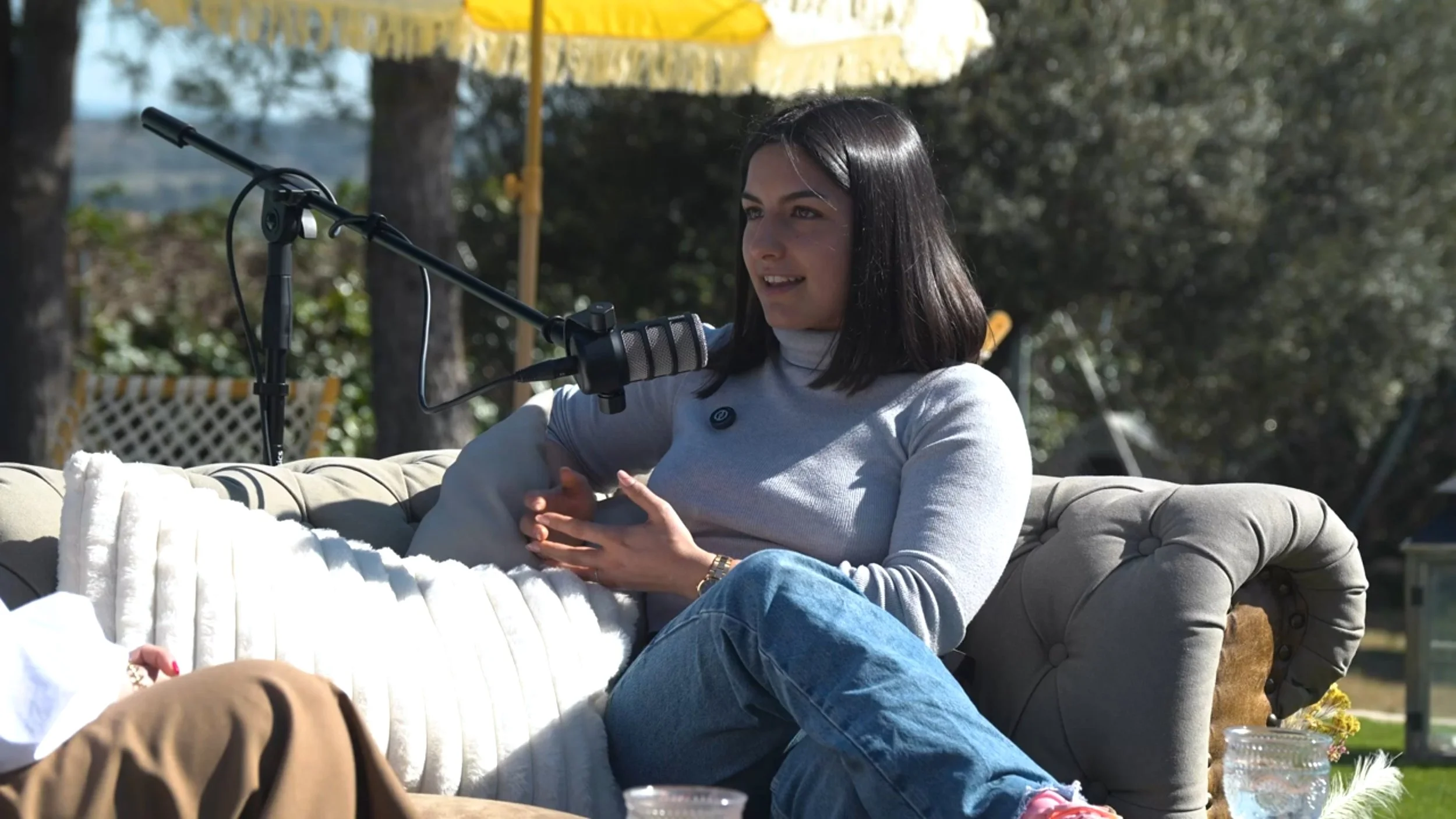 Una mujer sentada en un sofá al aire libre hablando en un micrófono durante una grabación o entrevista, con árboles y sombrillas de fondo.
