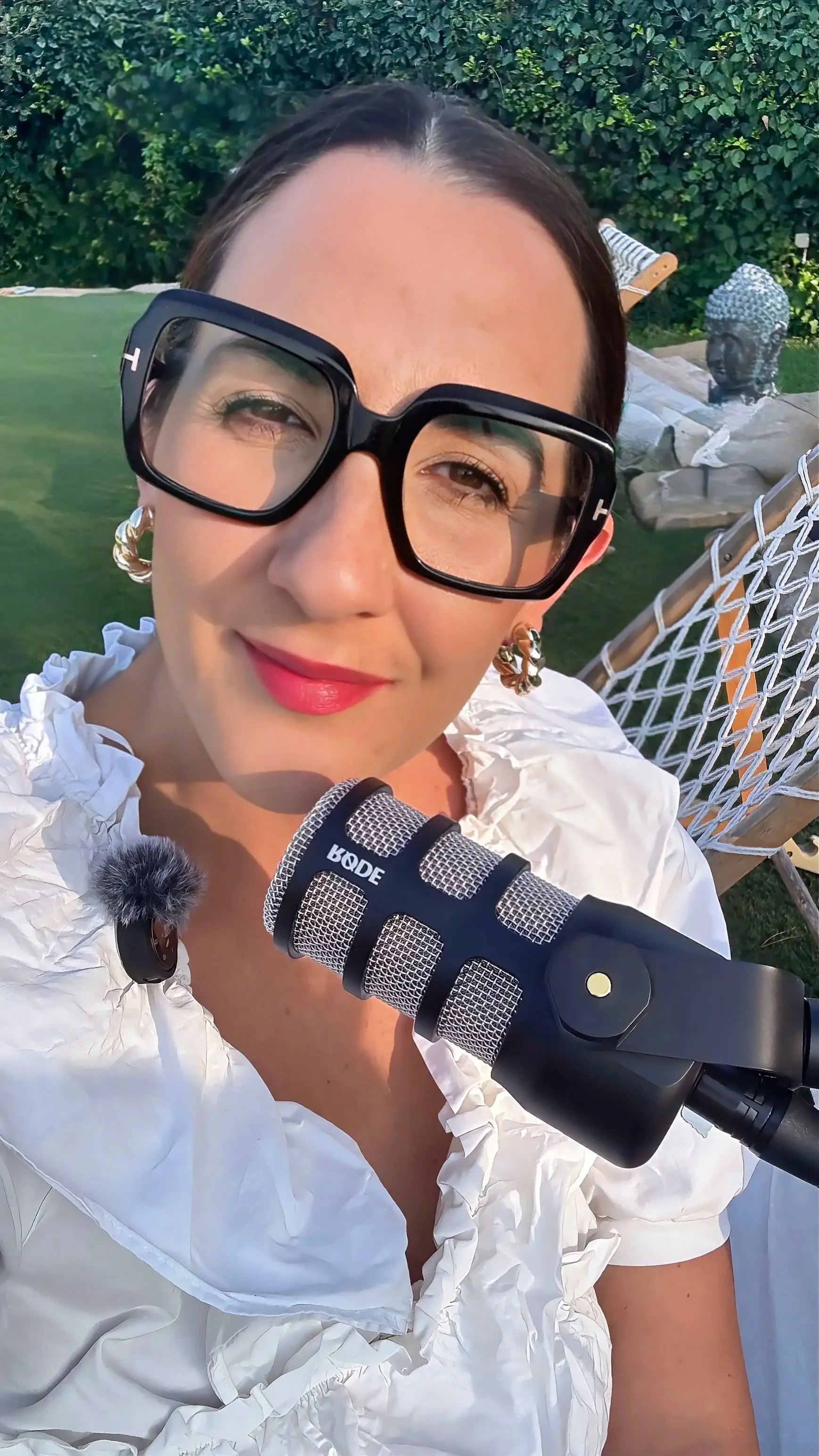 Mujer sonriendo con gafas grandes, sosteniendo un micrófono y usando una blusa blanca con volantes en las mangas. Fondo de un jardín con una escultura de cabeza de Buda y una tumbona.