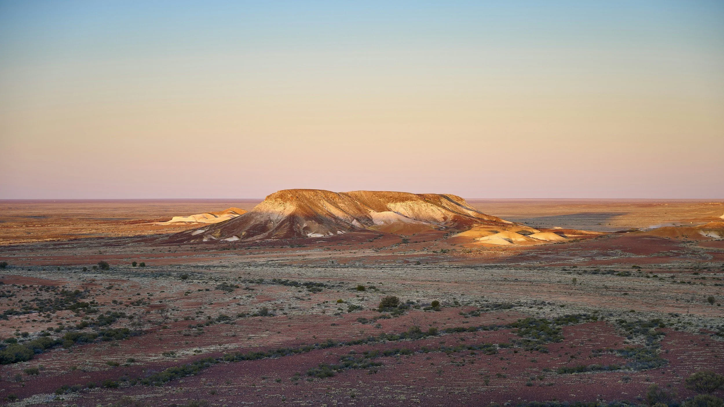 A desert landscape featuring a large, flat-topped mountain with colorful layers at sunset or sunrise, under a clear sky.