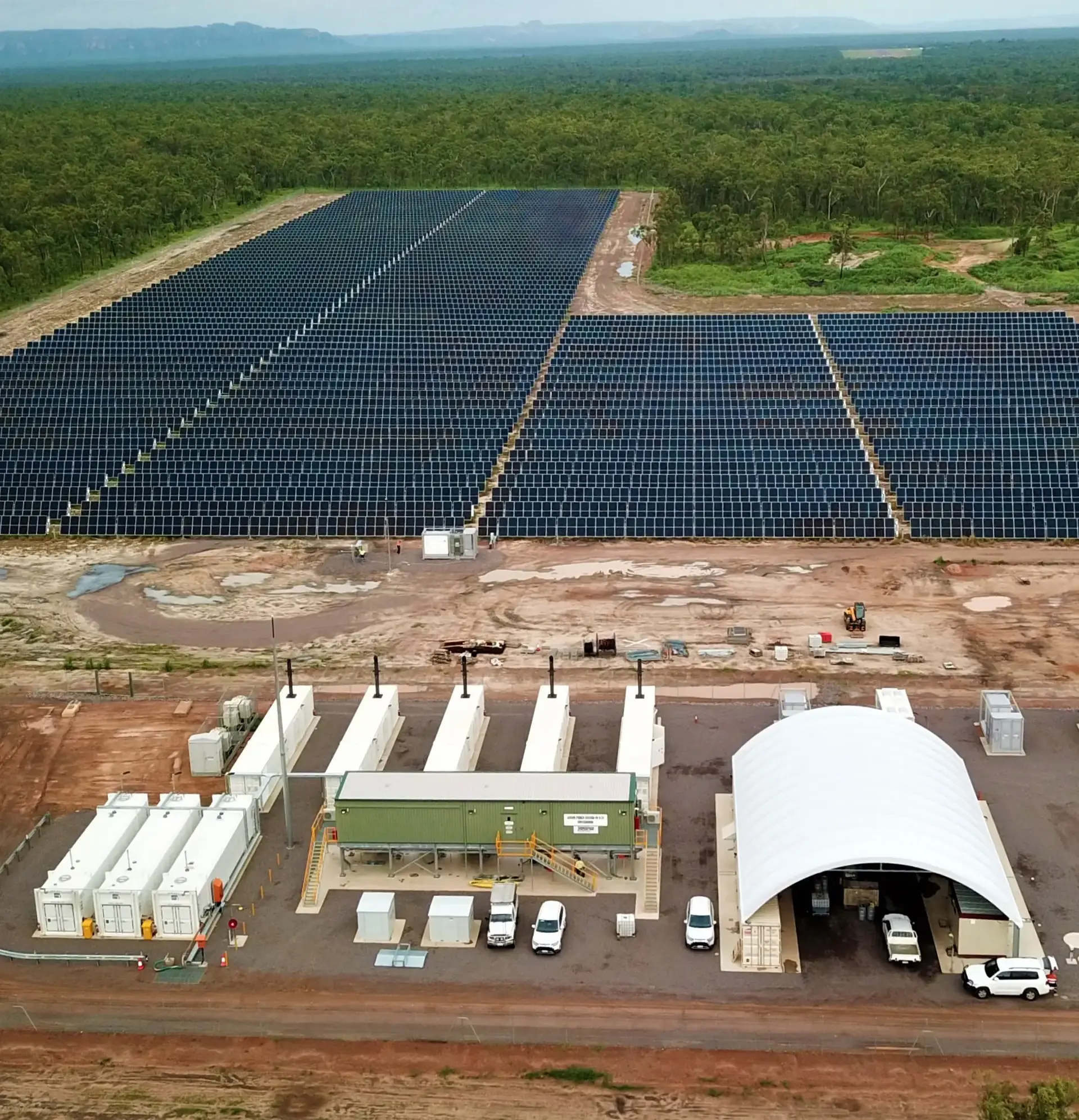 An aerial view of a large solar power facility surrounded by forest and dirt roads, with white modular buildings and parked vehicles in the foreground.