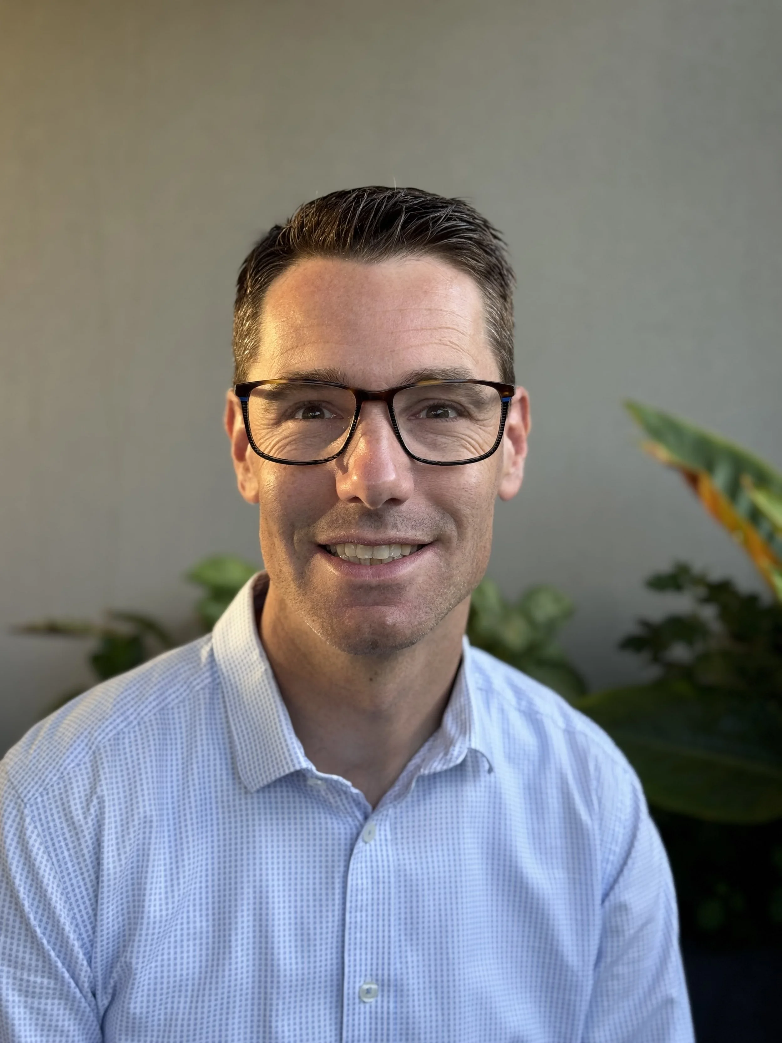 A smiling man with dark hair and glasses, wearing a light blue checkered shirt, standing outdoors with green plants and a gray wall in the background.