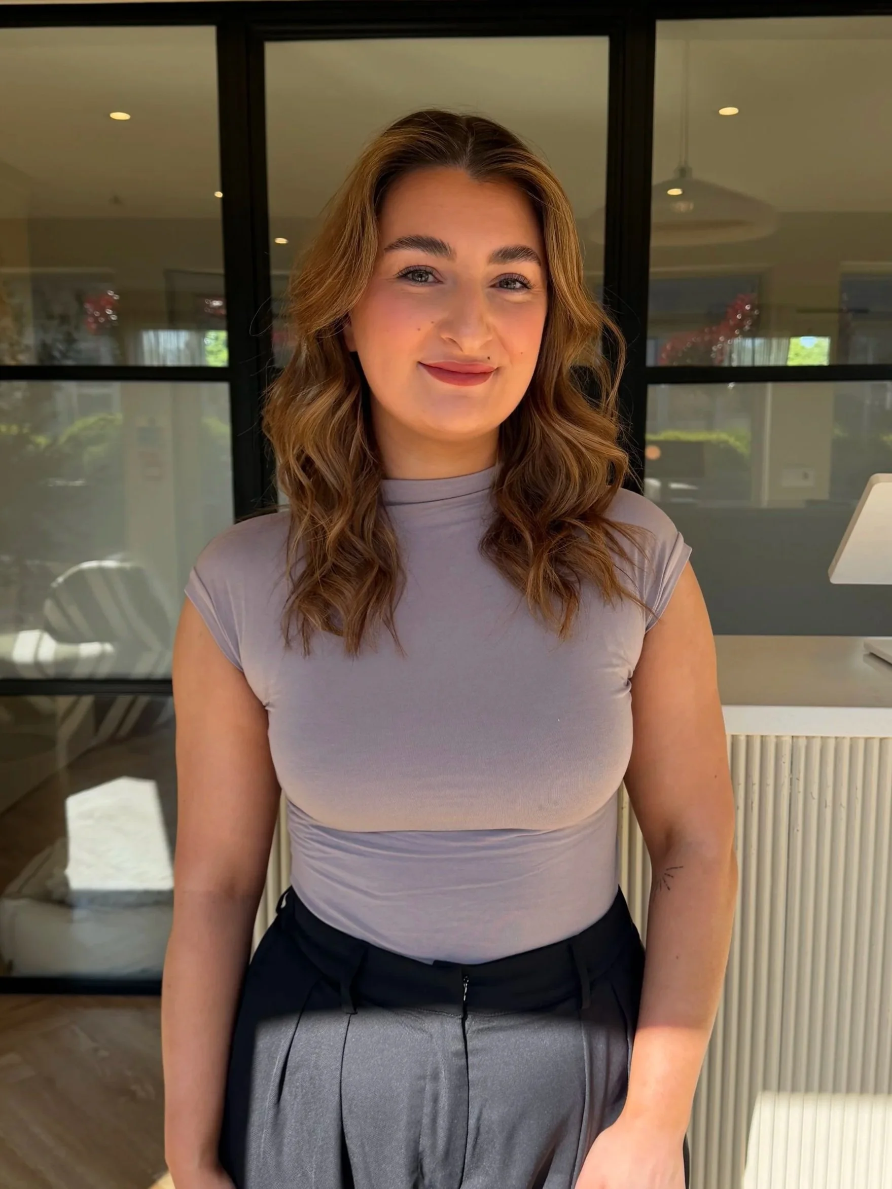A woman with wavy brown hair smiling and wearing a light purple top and gray pants standing indoors in front of glass doors.