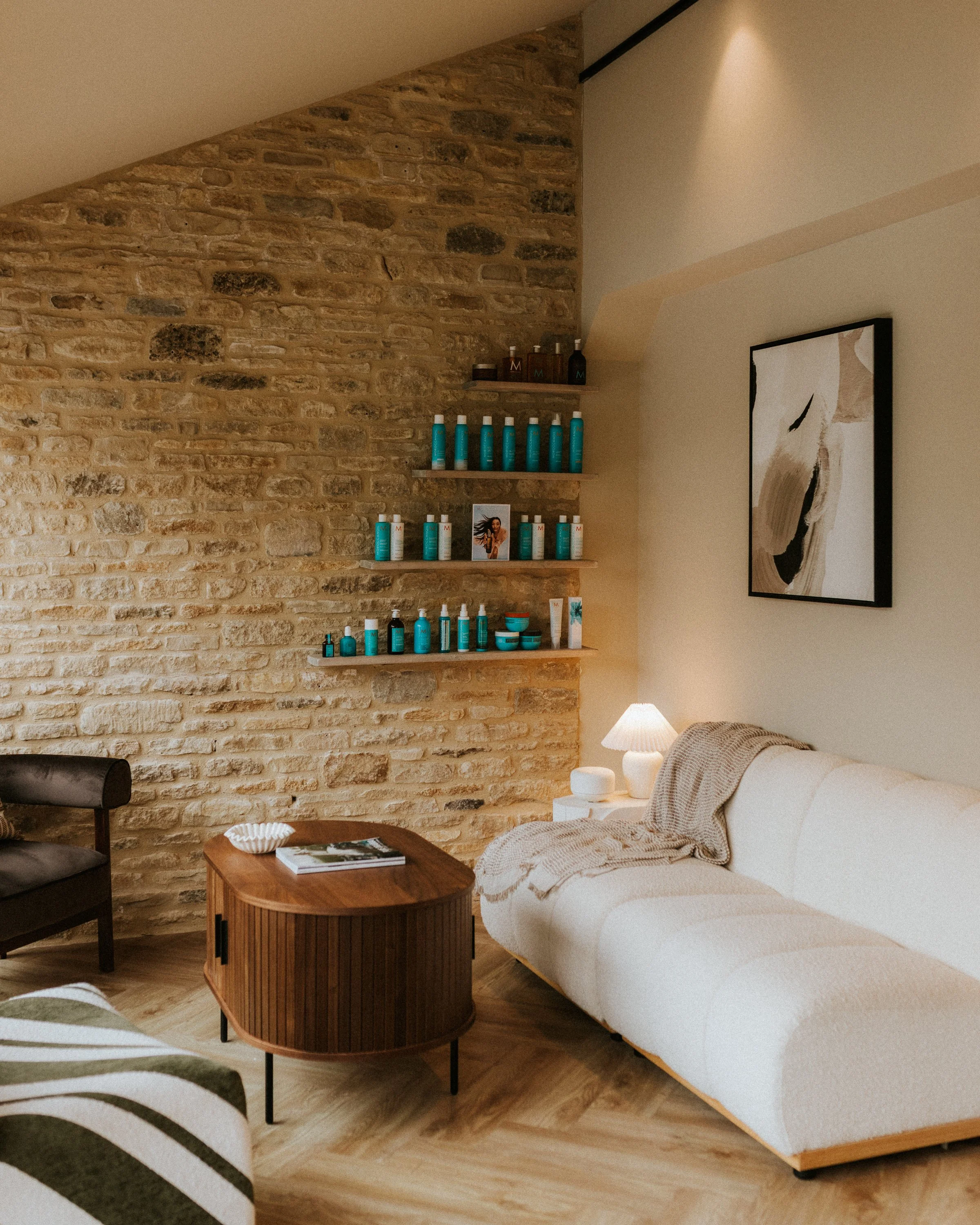 Living room corner with a white couch, wooden coffee table, and a brick accent wall with floating shelves holding skincare products. A framed abstract art piece hangs on the beige wall and a small lamp is on a side table.