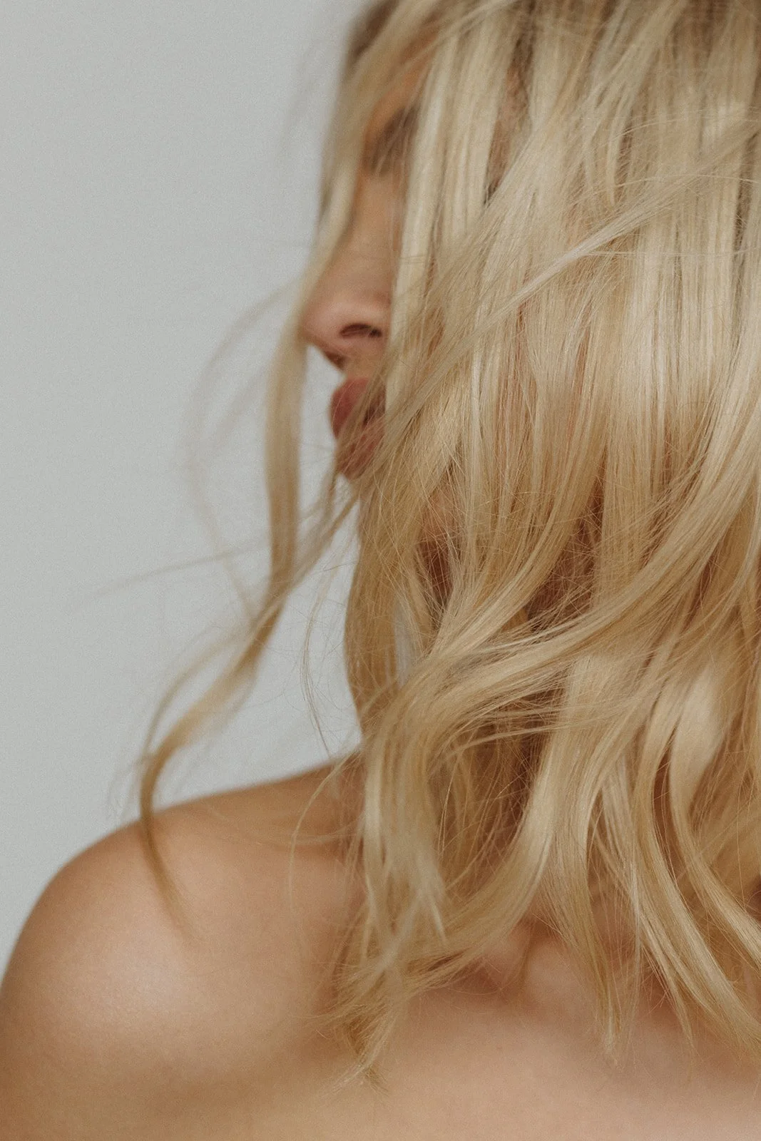 Close-up of a woman with long, wavy blonde hair, partially covering her face, showing her nose, lips, and shoulder against a plain, light background.