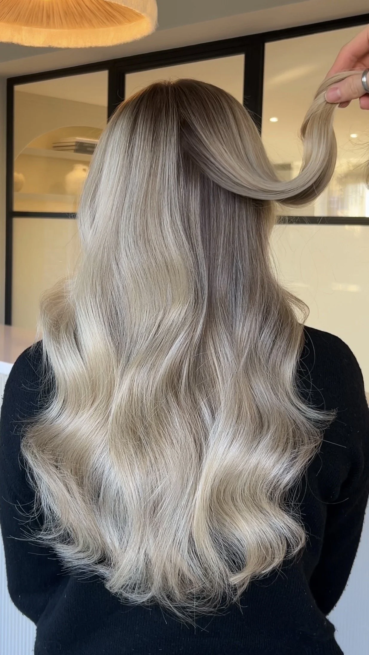Back view of a woman with long, wavy, blonde hair with darker roots, in a salon or studio setting.