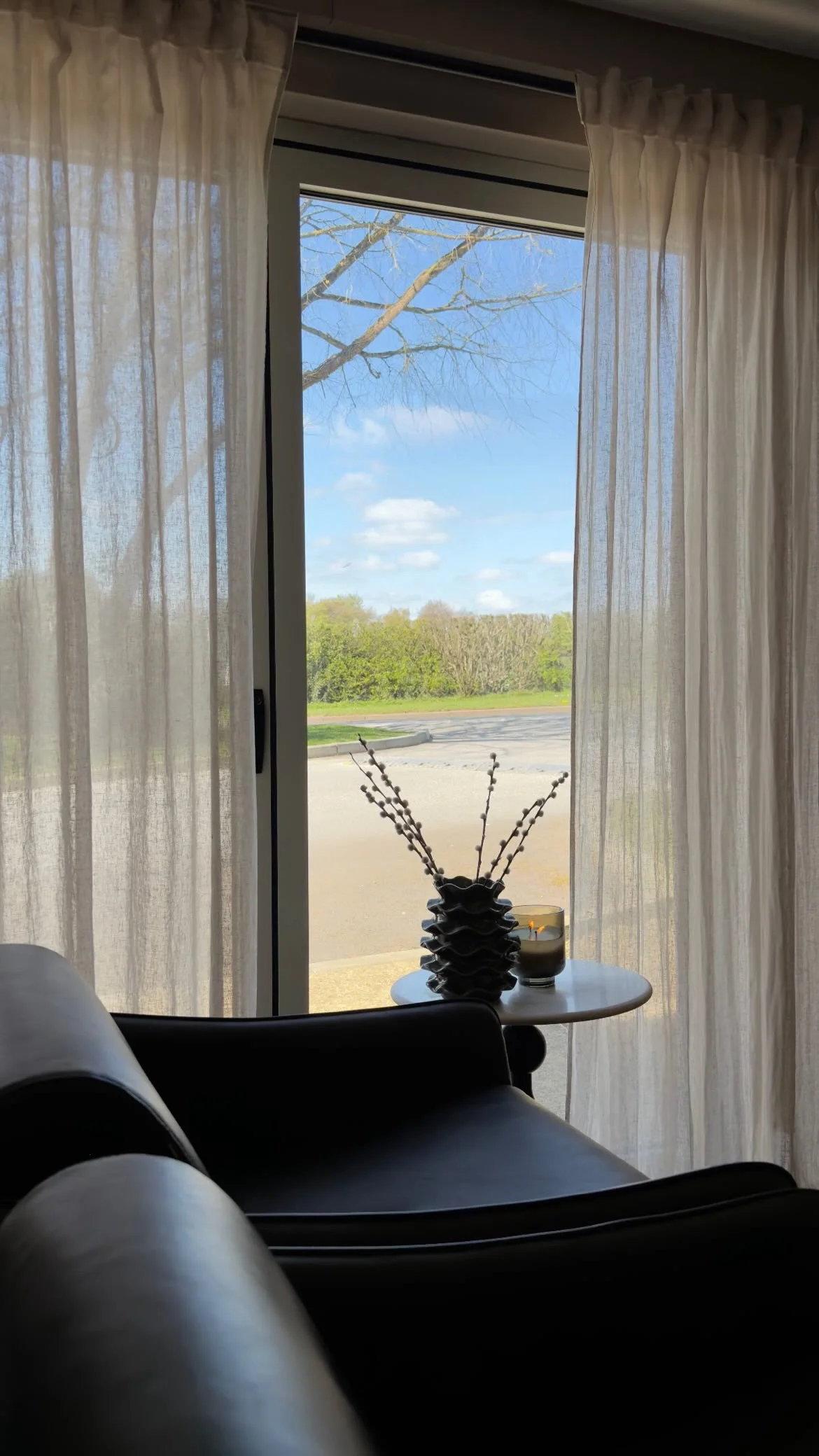 View through a sliding glass door with sheer white curtains, showing a clear blue sky, leafless tree branches, and green trees in the distance. Inside, there is a small white table with a decorative vase holding twigs and a candle.