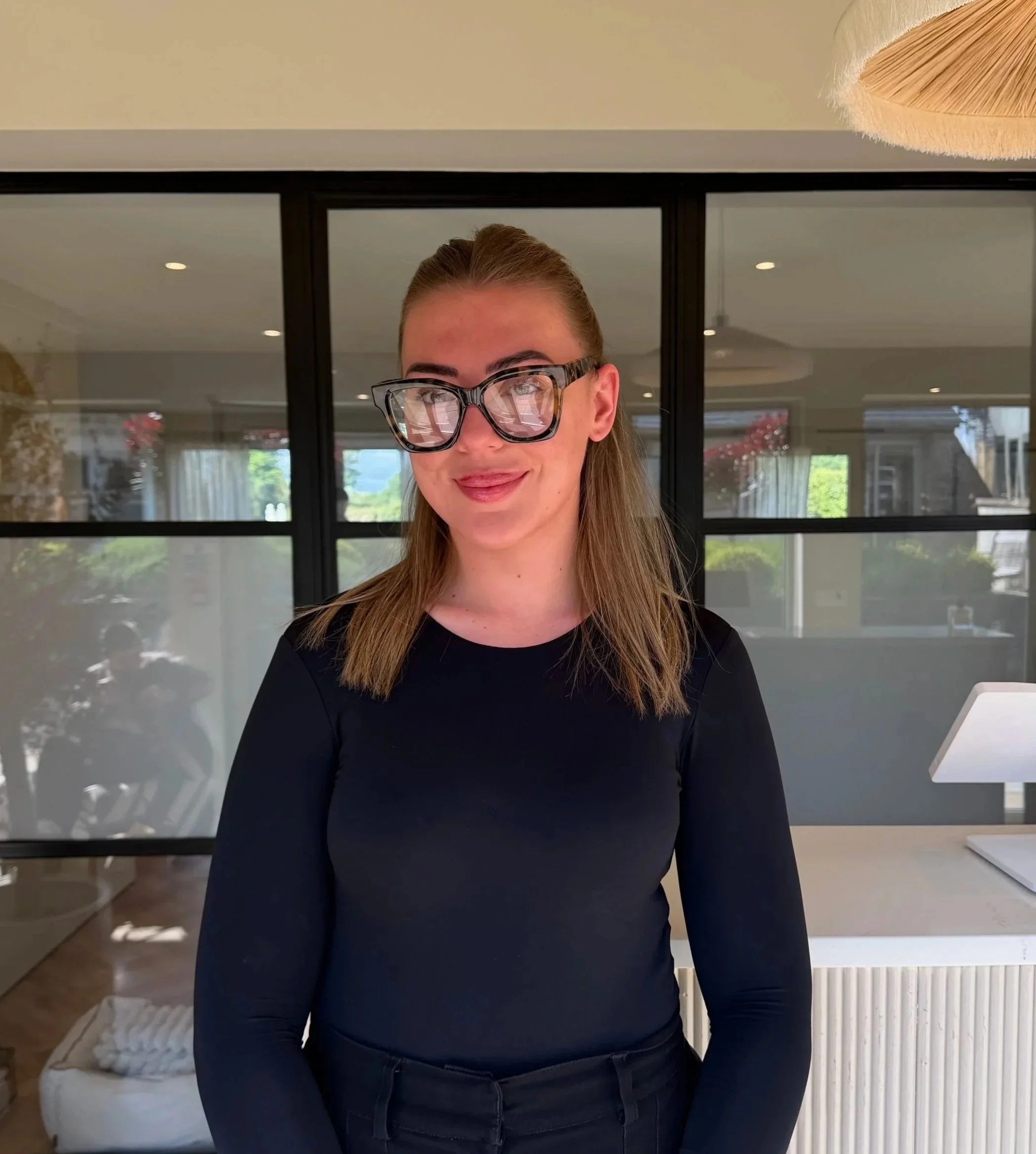 Young woman with long light brown hair, wearing black glasses and a black top, smiling in a modern room with large window and indoor plants.