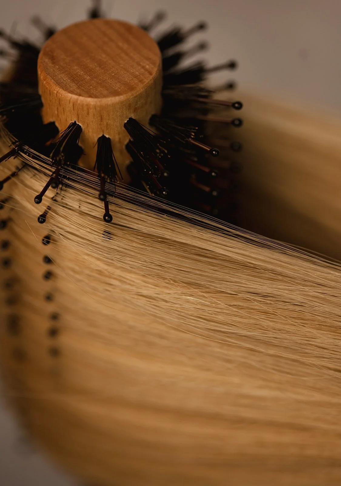 Close-up of a round hairbrush with a wooden handle, black bristles, and light blonde hair strands.