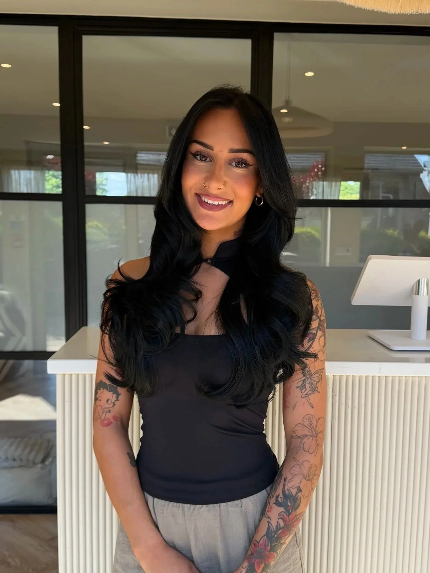 A woman with long black hair, tattoos on her arms, and wearing a black sleeveless top is smiling at the camera inside a modern building with large windows and minimalist decor.