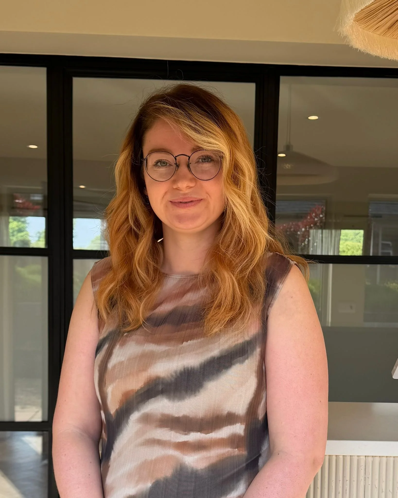 A woman with red hair, glasses, and a sleeveless dress standing indoors with large glass windows behind her, showing greenery outside.