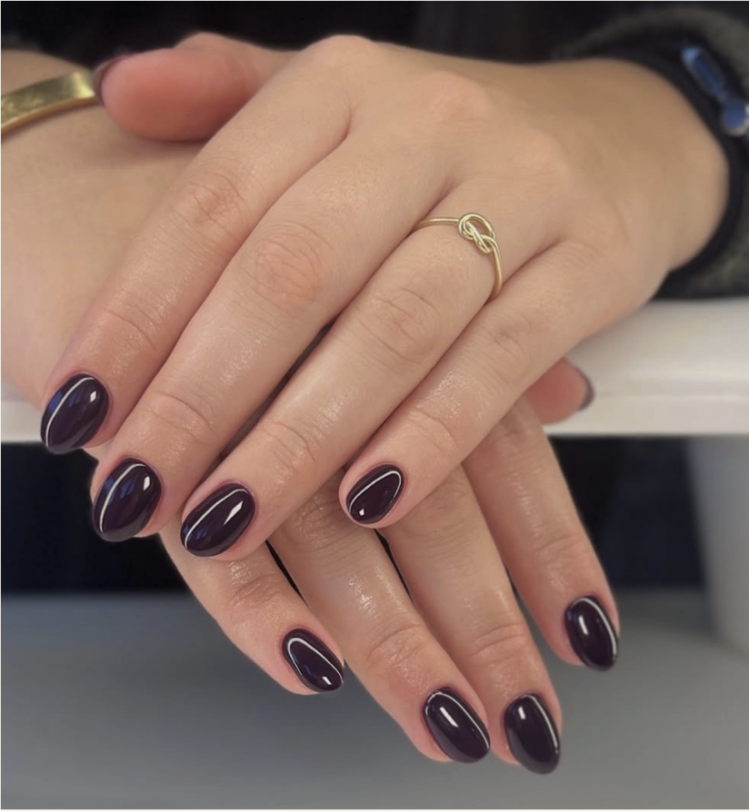 Close-up of a person's hands with manicured nails painted dark purple with light purple tips, displaying a gold infinity ring on the index finger and a gold bracelet on the wrist.