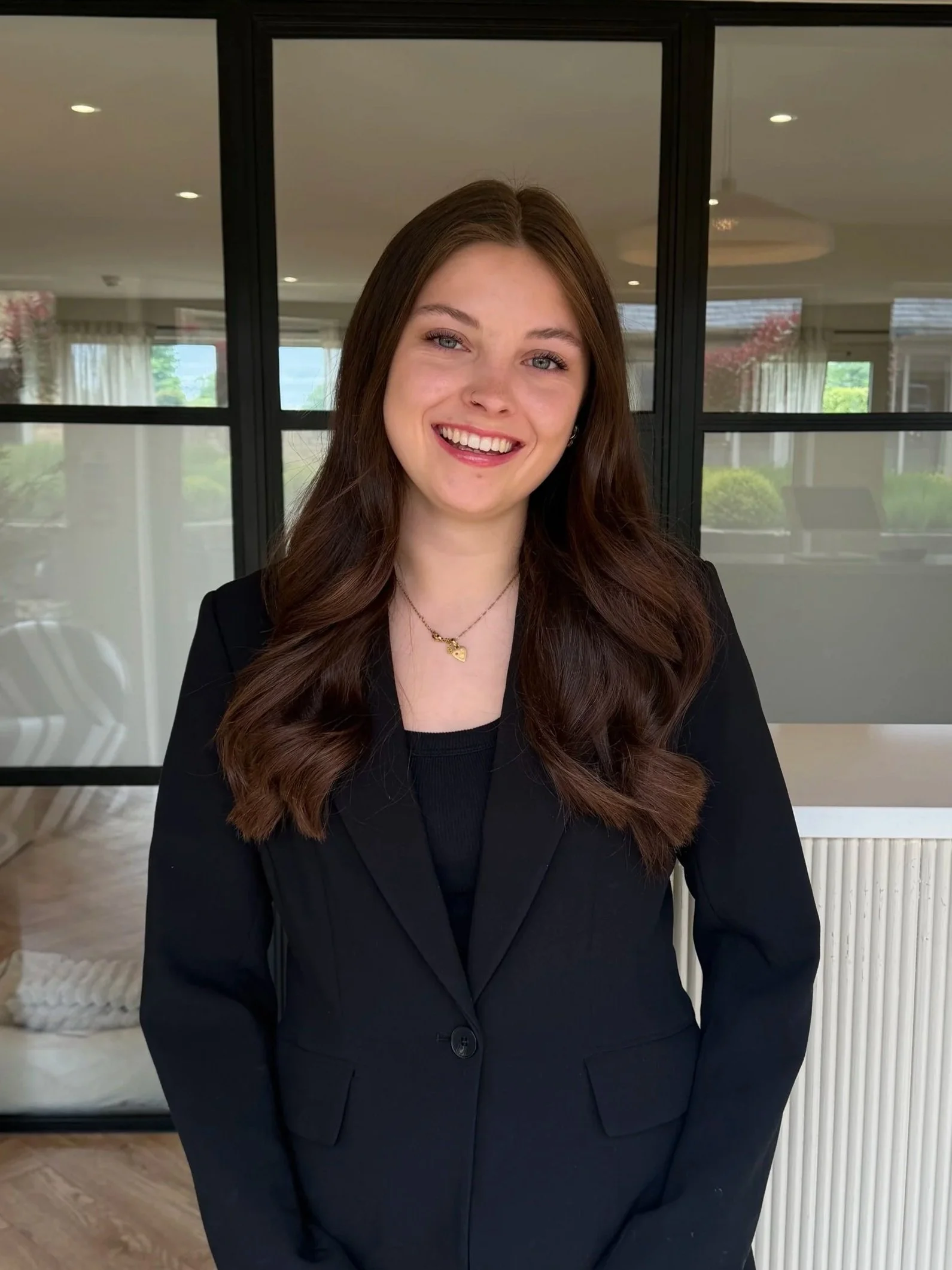 A smiling woman with long brown hair, wearing a black blazer and a gold heart necklace, sitting indoors with a modern background of large glass panels and a white radiator.
