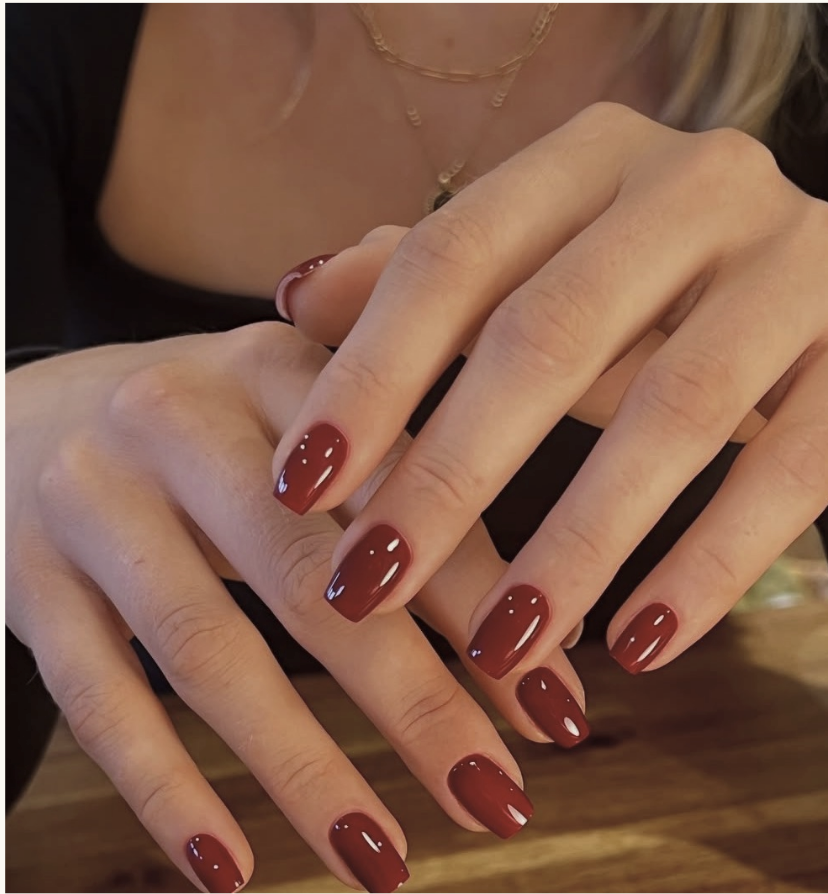 Close-up of hands with red painted nails, adorned with gold rings and layered delicate necklaces, resting on a wooden surface.