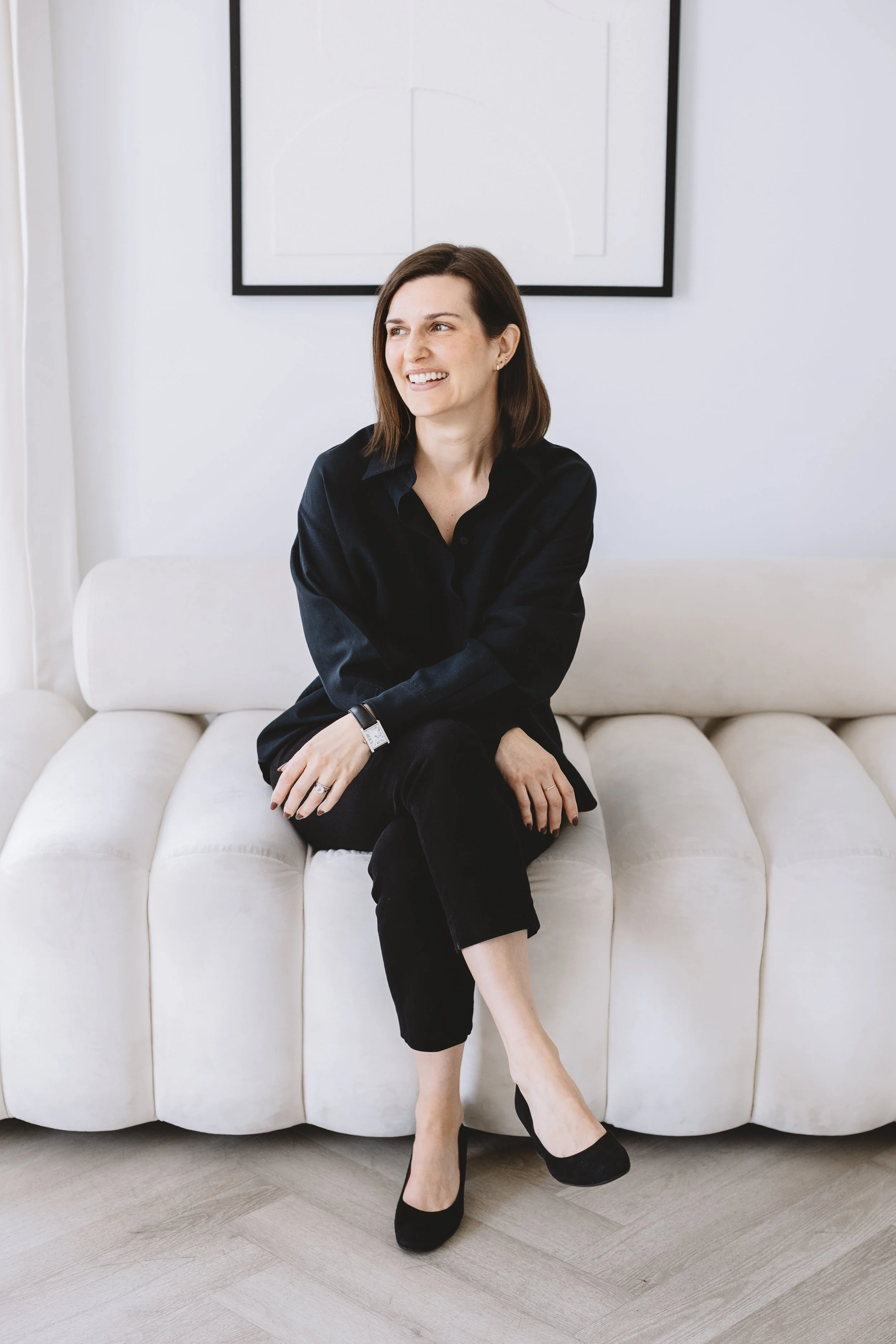 A woman with brown hair wearing a black shirt and pants sitting on a white sofa, crossing one leg over the other, smiling, in a modern room with a white wall and framed black and white artwork behind her.