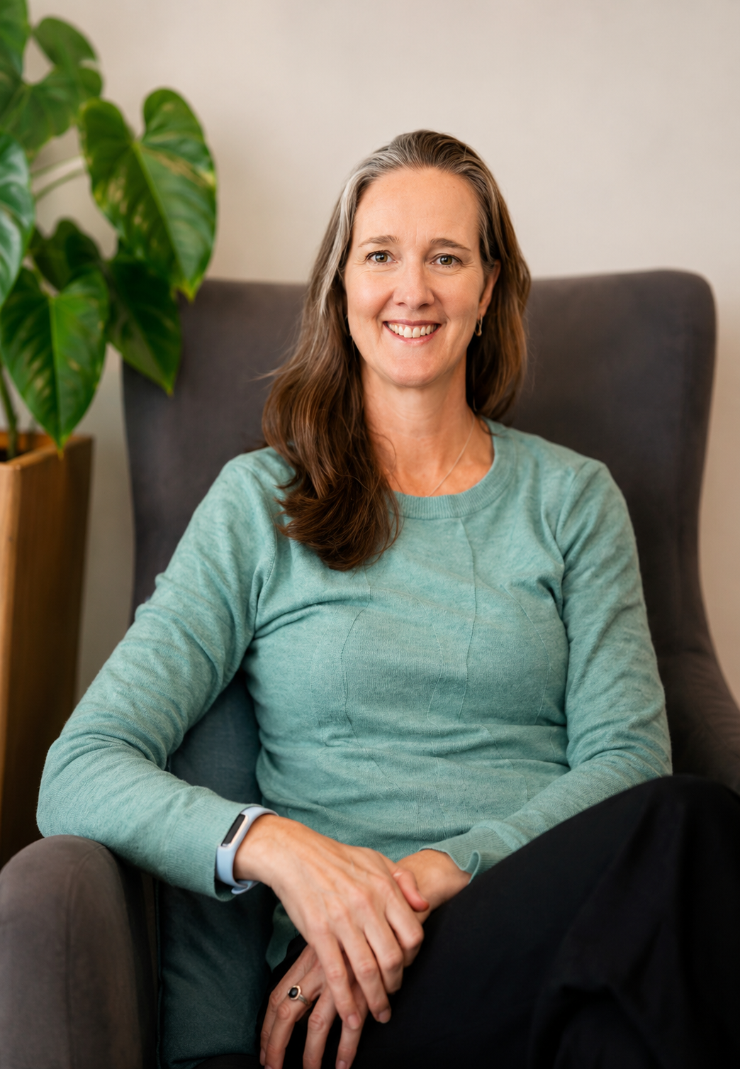 A woman with brown hair, wearing a light green sweater, seated comfortably in a gray armchair, smiling with a houseplant in the background.