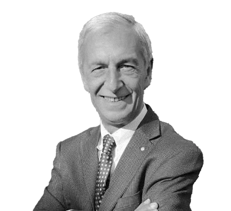 Un uomo anziano con capelli bianchi, indossa un abito grigio con una camicia bianca e una cravatta con pois, sorridente, con le braccia incrociate.