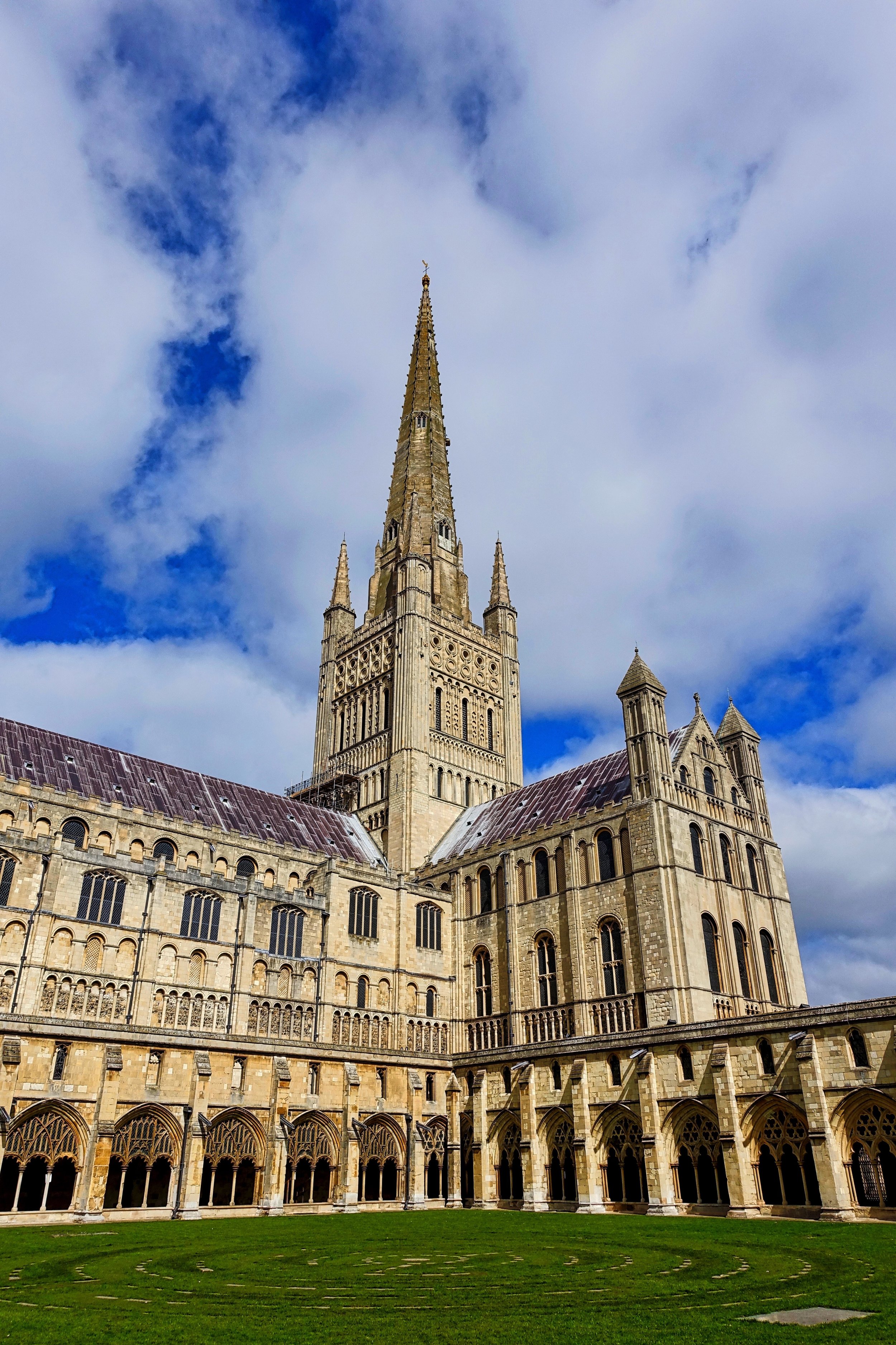 Norwich cathedral with a tall, pointed spire reaching into a partly cloudy sky, surrounded by a green lawn.