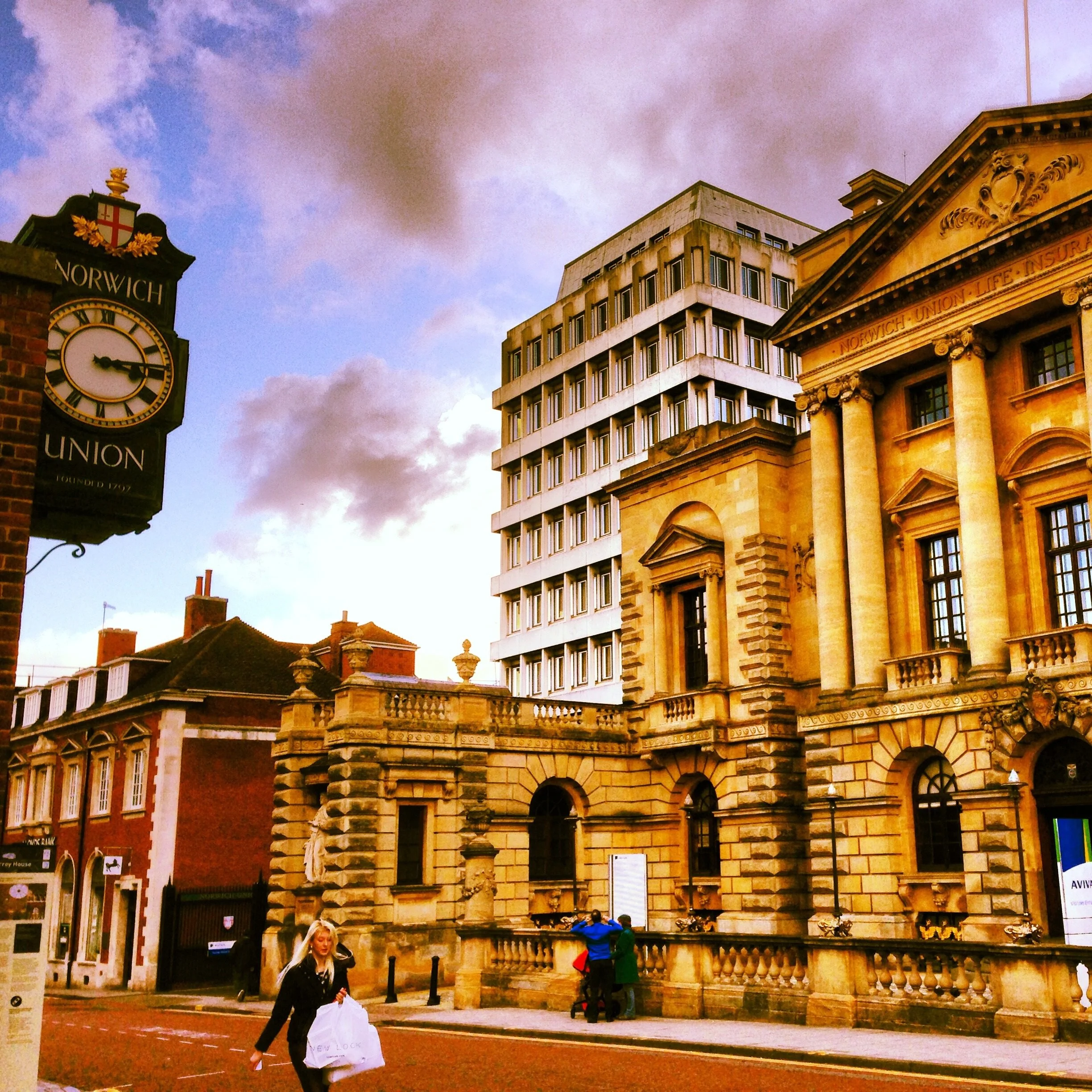 Street scene with historic and modern buildings, a clock tower with a gold crest reading "Norwich" and "Founded 1797," and people walking along the sidewalk during dusk. The sky shows a mix of blue and pink clouds.