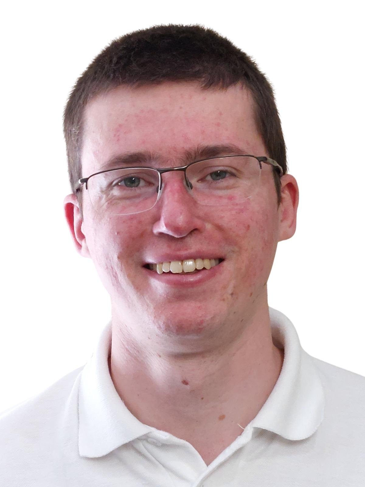 Portrait of a smiling young man with glasses, short dark hair, and light skin, wearing a white collared shirt against a white background.