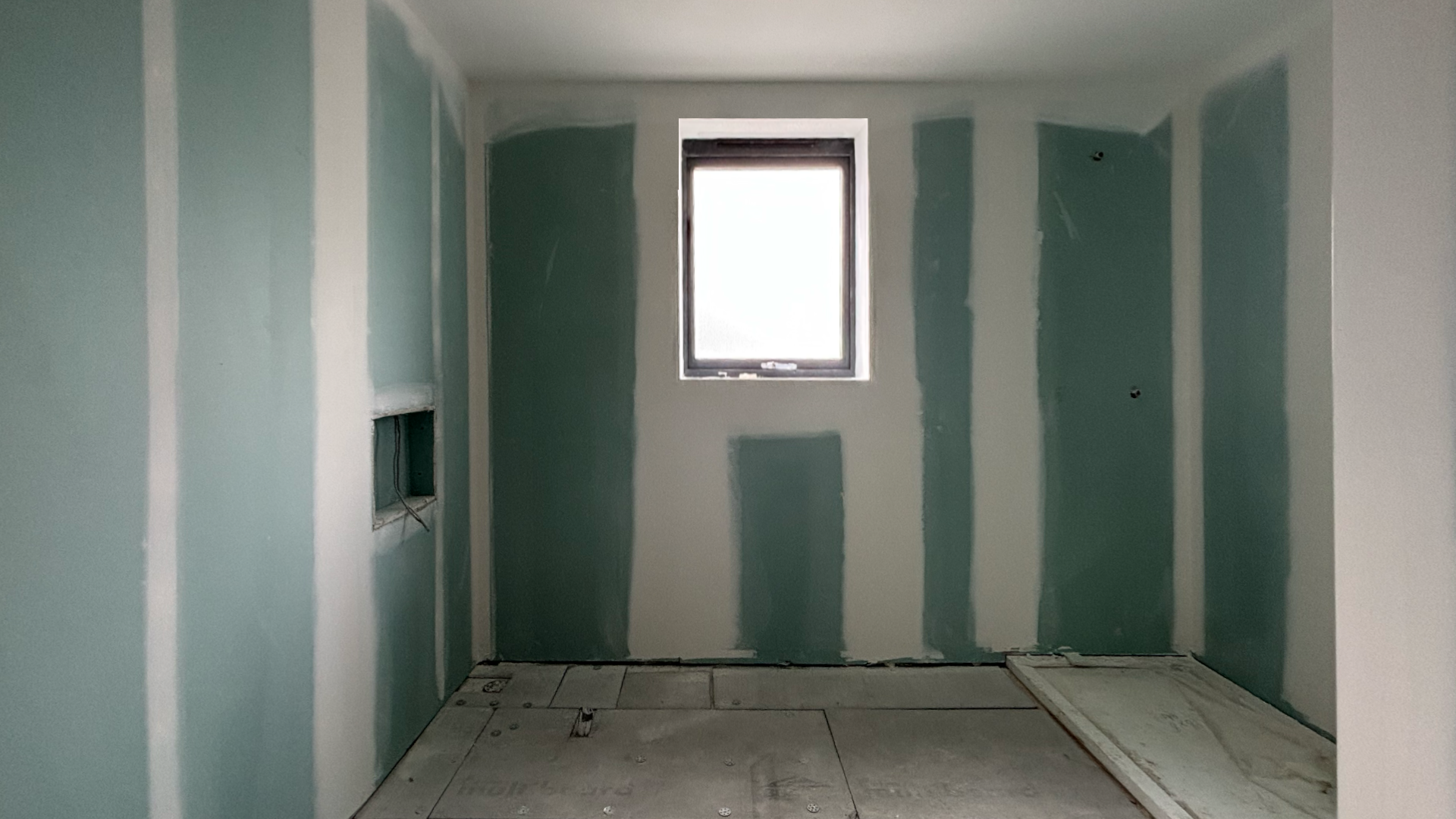 An unfinished room with drywall installation, featuring a window, electrical wiring, and flooring under construction.
