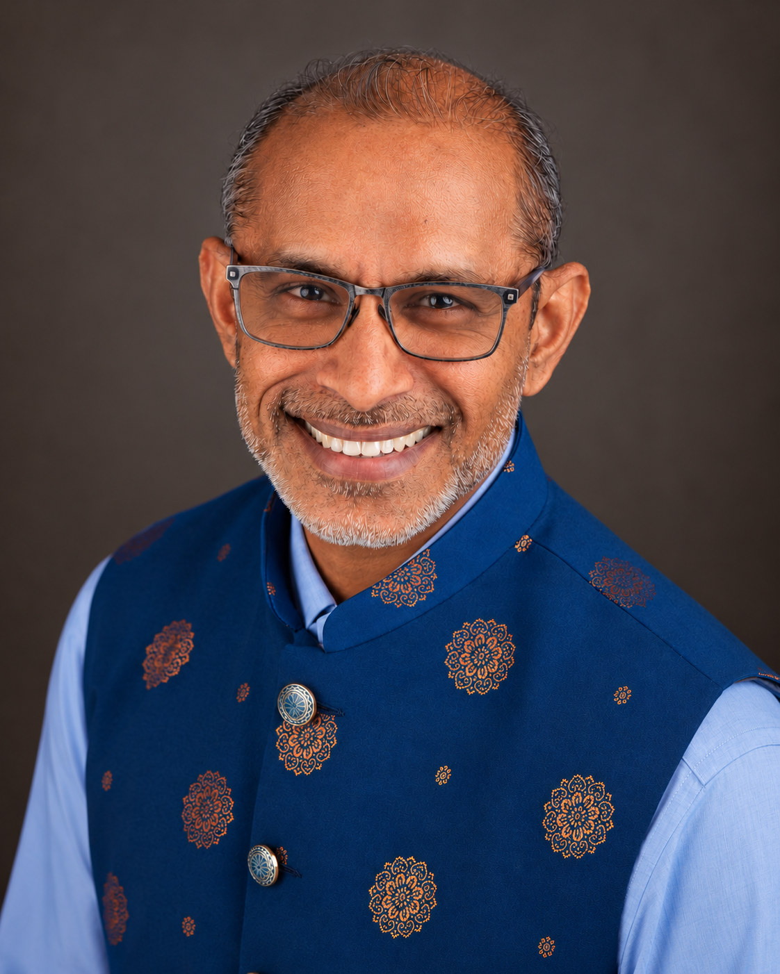 Professional headshot of a smiling man in a blue embroidered vest and glasses.