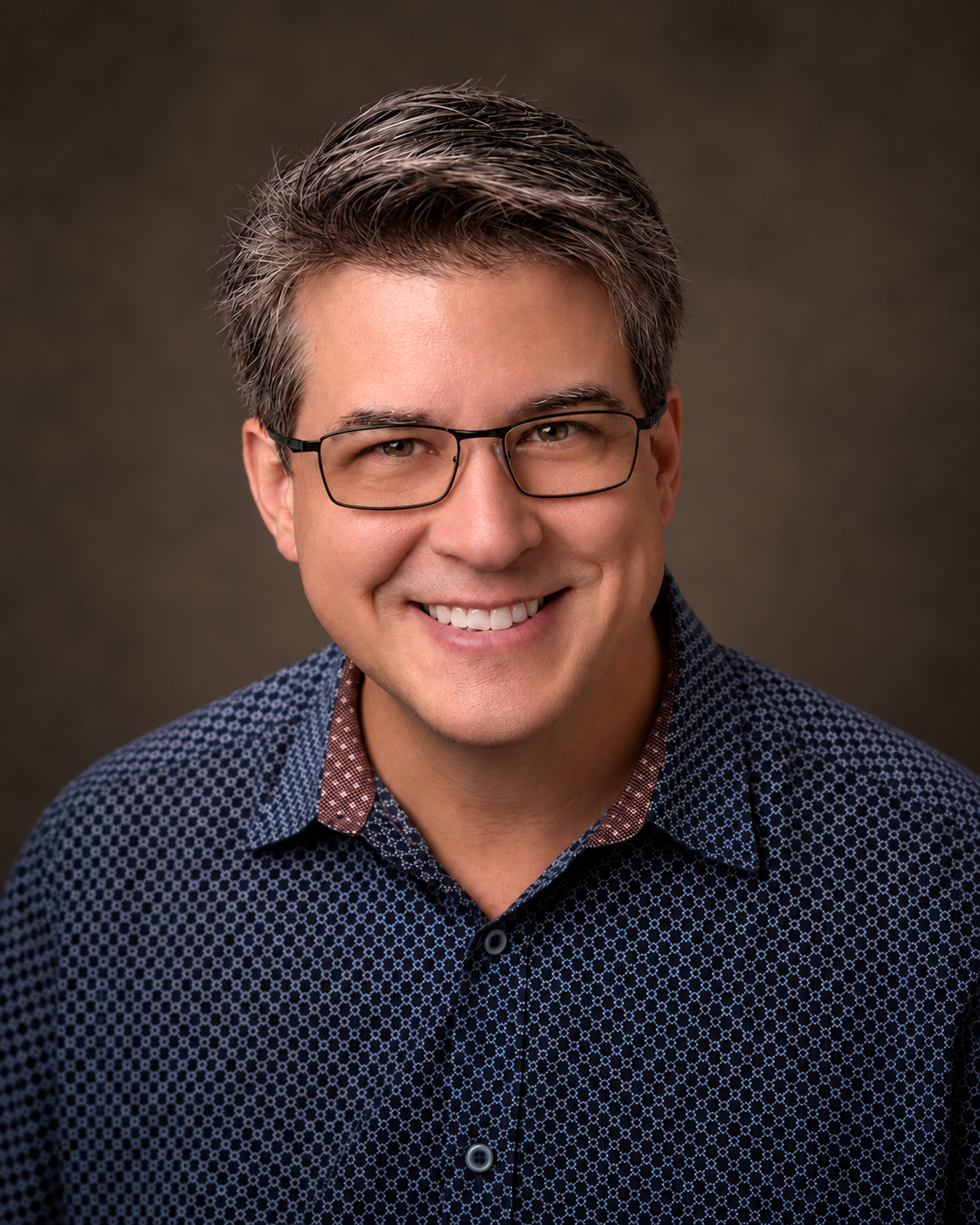 Professional studio headshot of a smiling man wearing glasses and a patterned blue shirt.