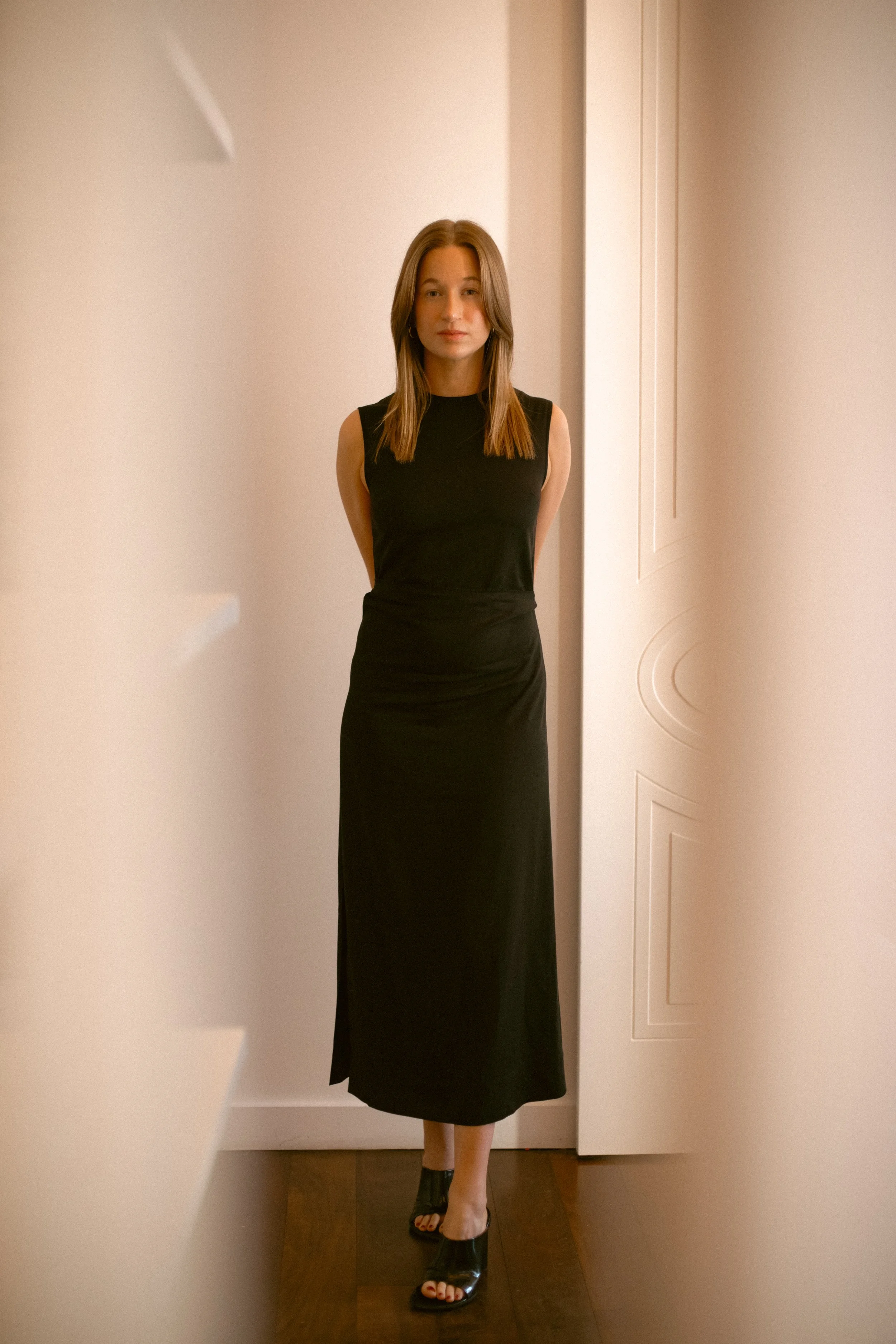 A woman stands in a room with her hands behind her back, wearing a sleeveless black dress and black open-toe heels, with a white door and beige walls in the background.