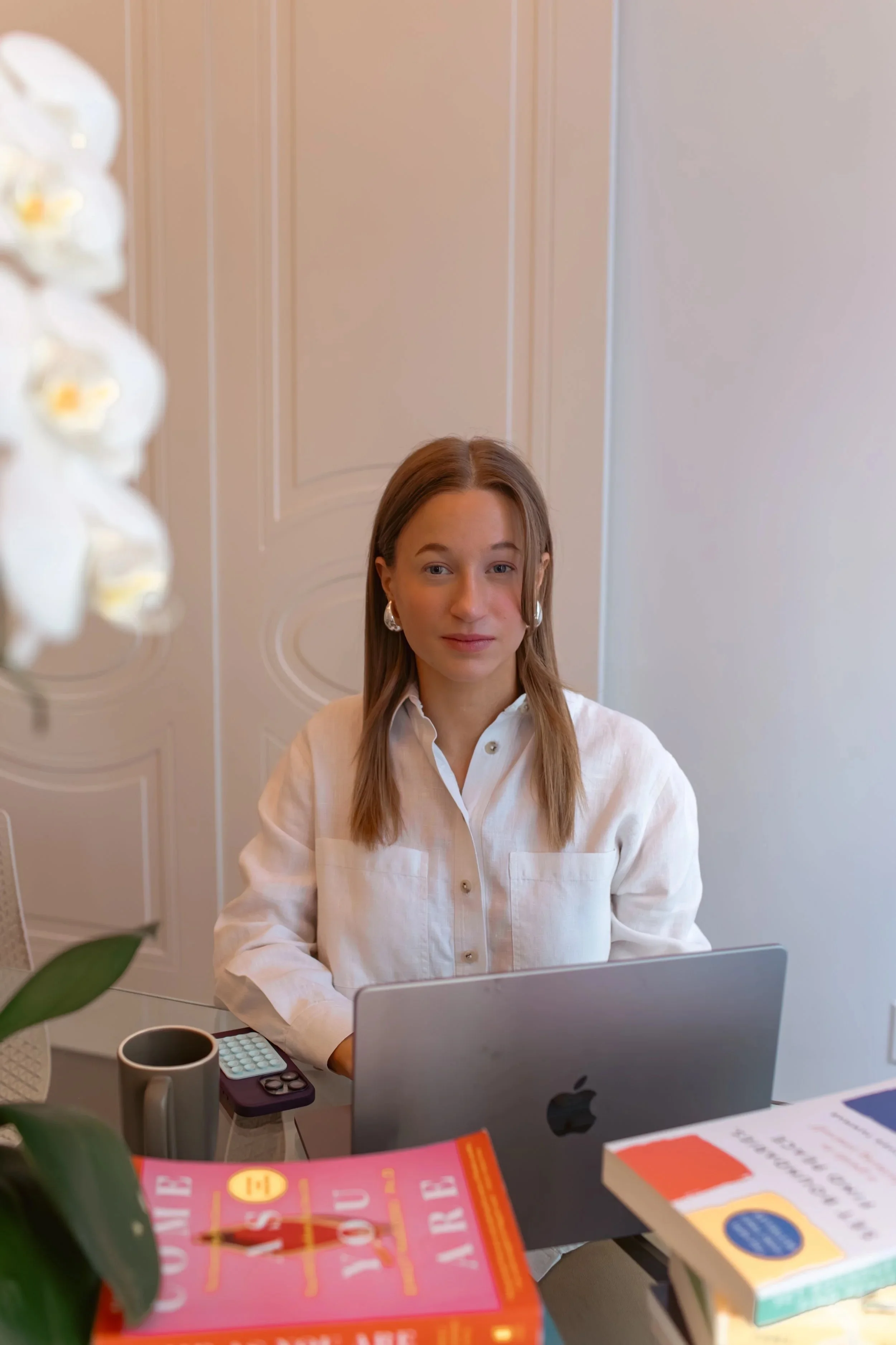 A woman with long brown hair and dangling earrings sitting at a desk with a laptop in an office or home setting. There are books, a plant, and a mug on the desk.
