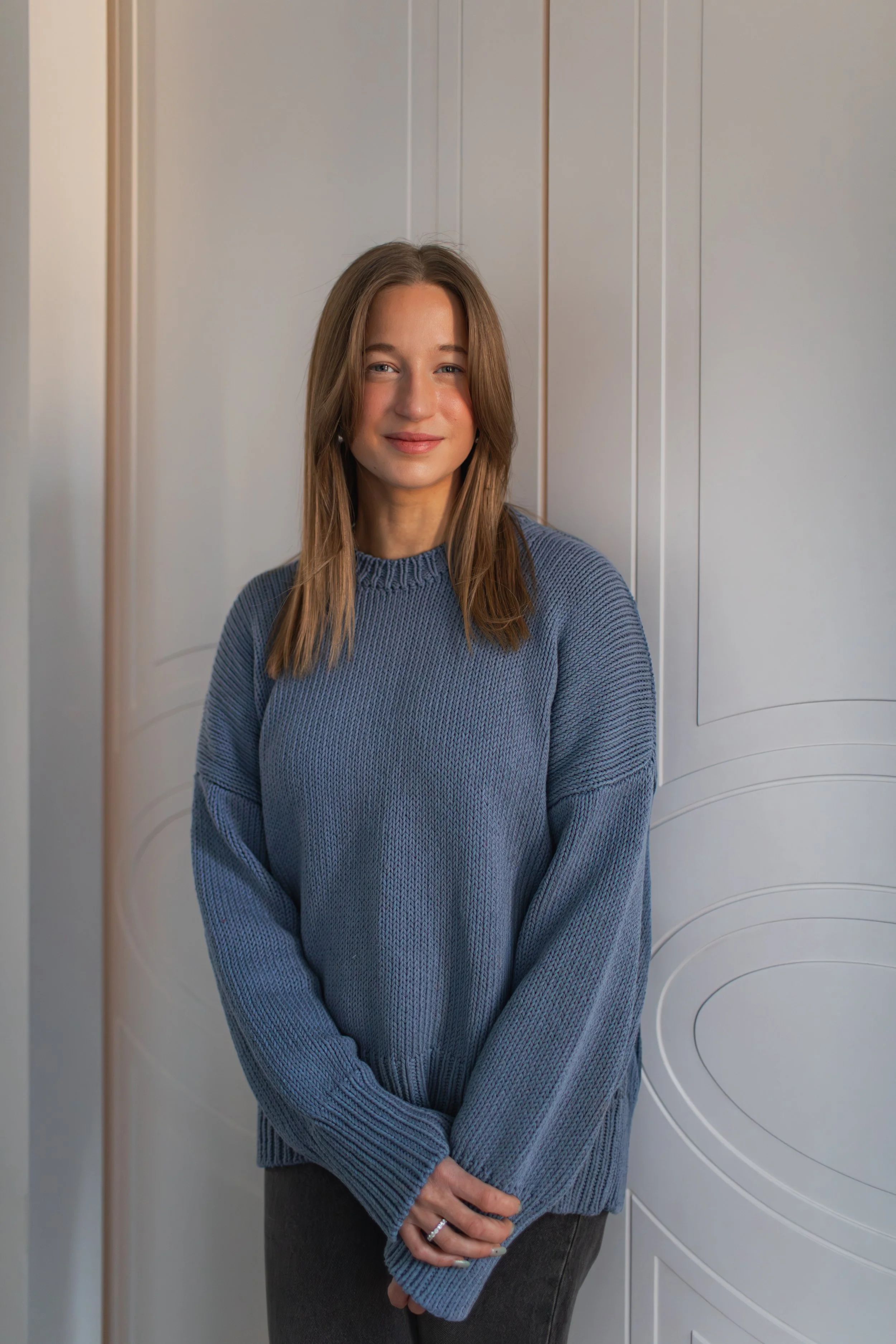 A woman with shoulder-length brown hair and blue eyes standing with arms crossed in front of a white door with curved paneling, wearing a blue knitted sweater and black pants.