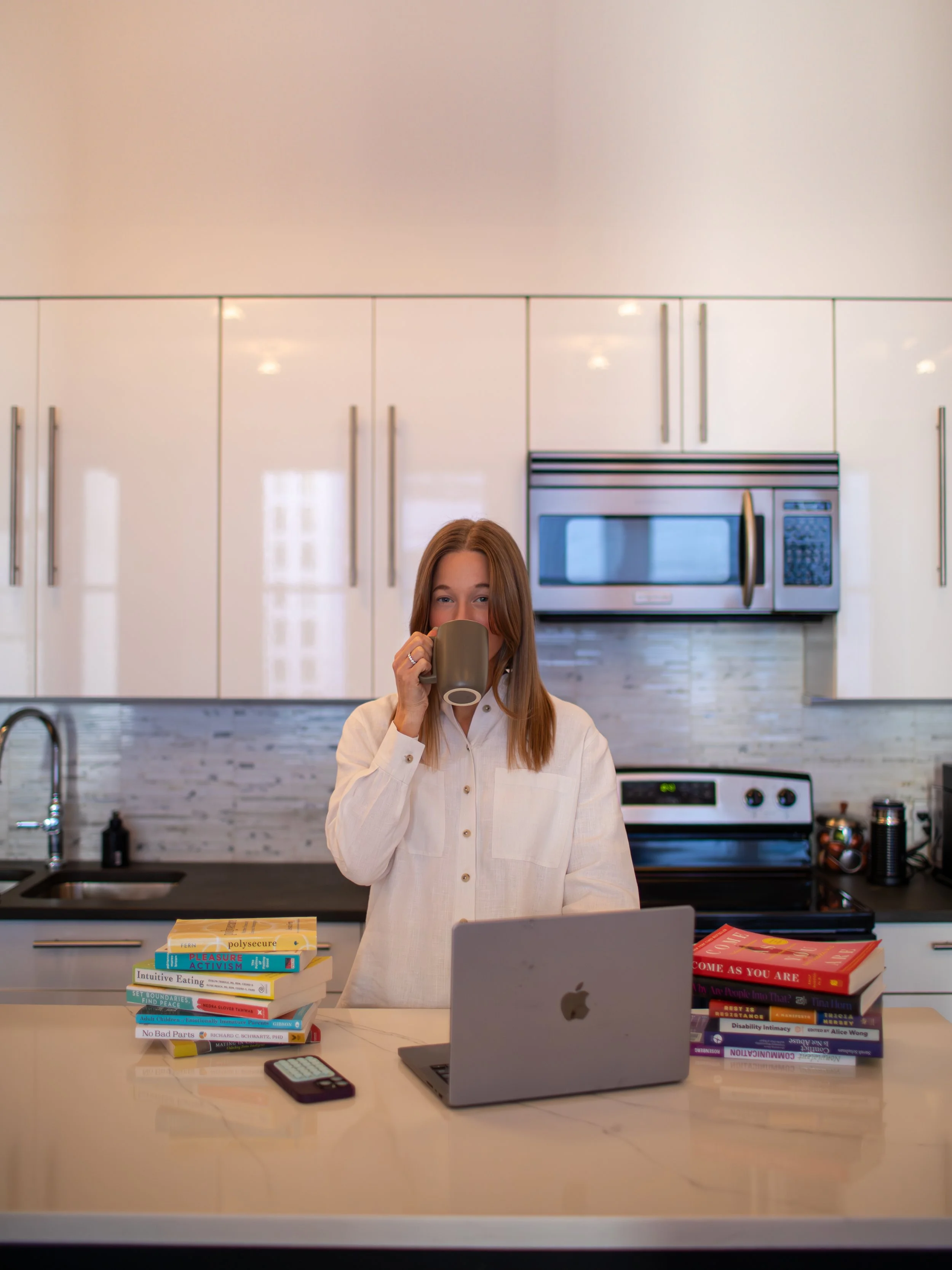 A woman with red hair drinking from a mug in a kitchen, with two stacks of books on the counter and an open laptop in front of her.
